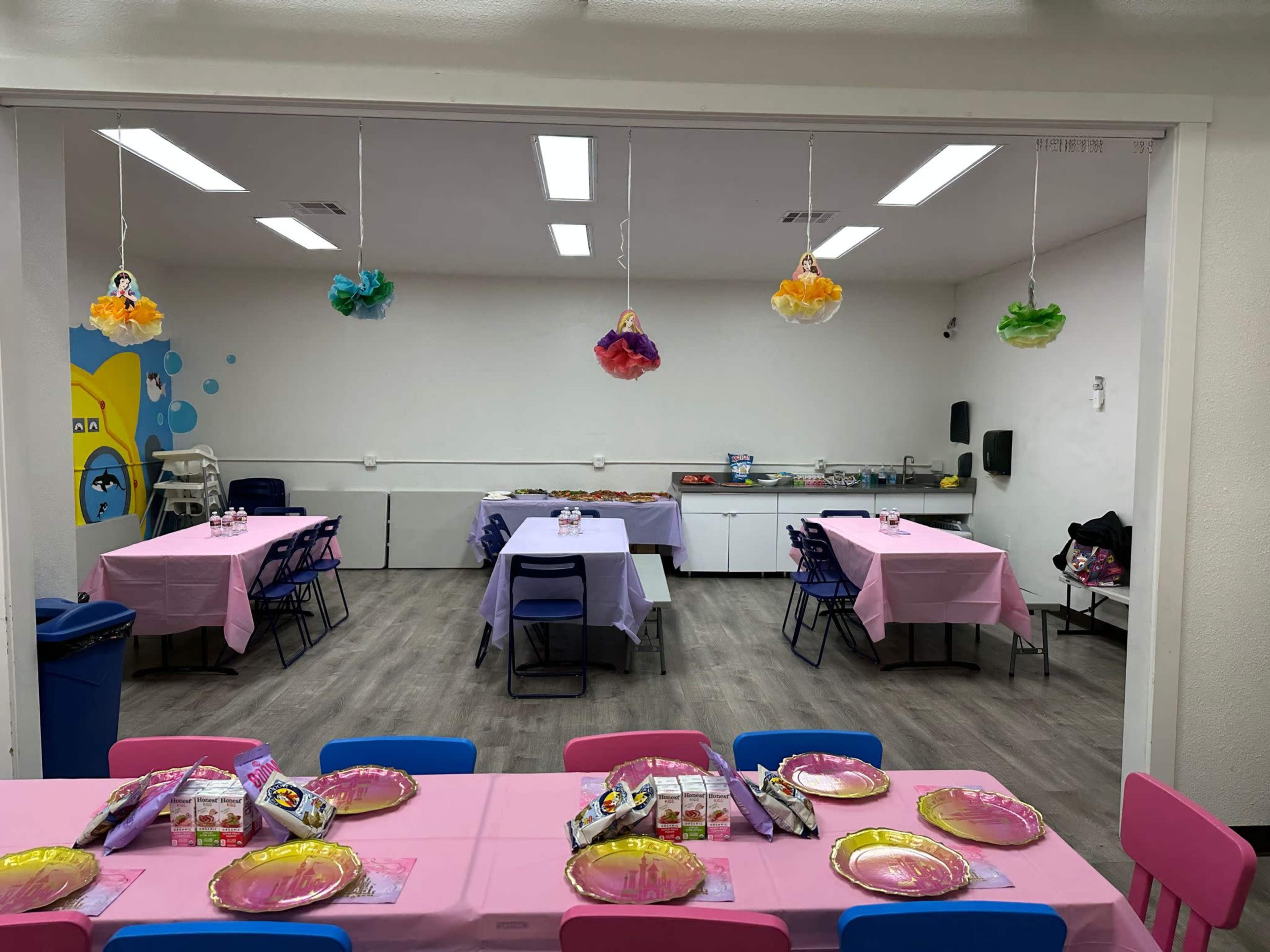 A colorful party room with tables covered in pink tablecloths and decorated with paper flowers hanging from the ceiling.