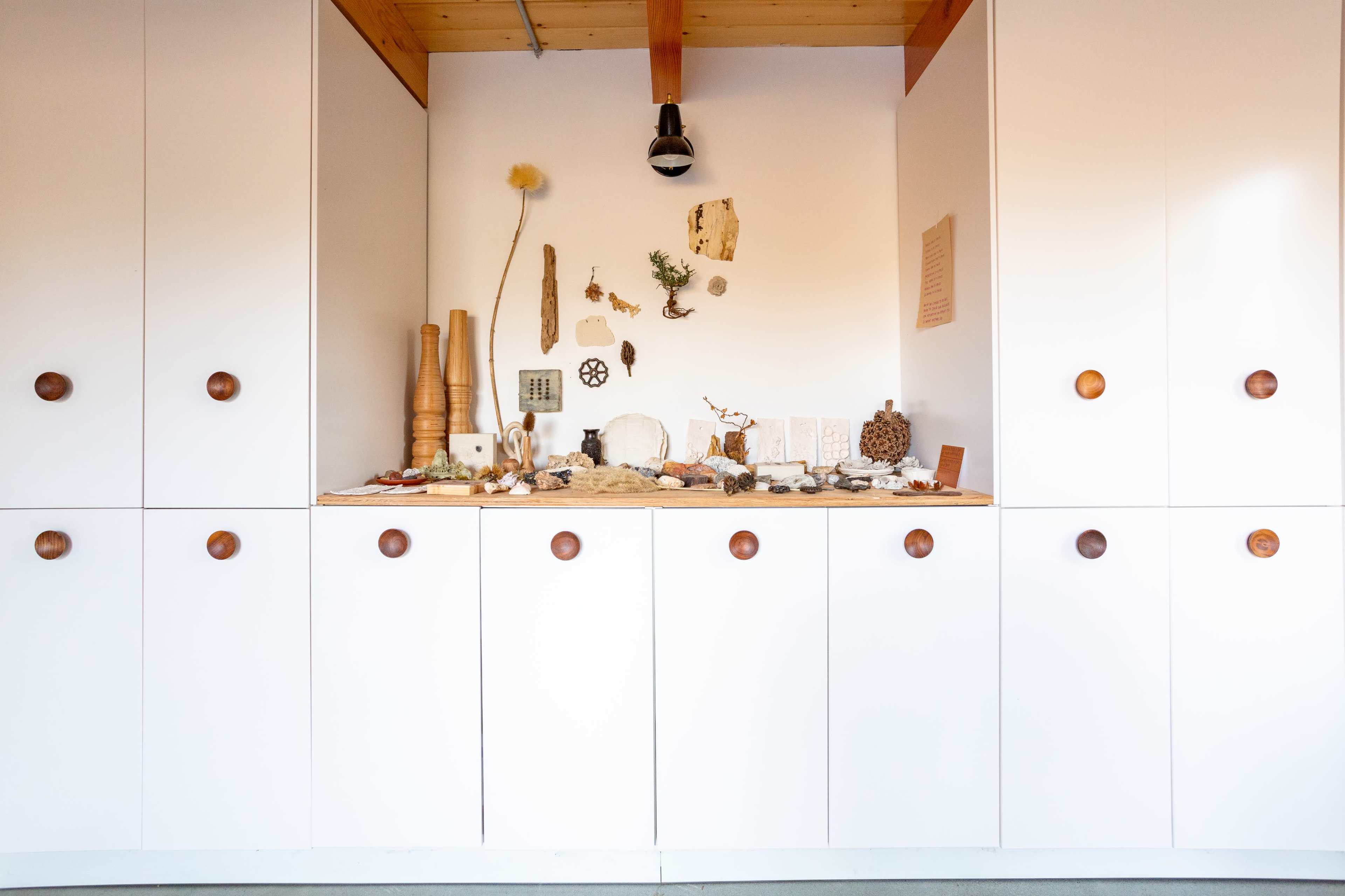 A wooden shelf displays a collection of decorative objects and natural materials against a backdrop of white cabinetry.
