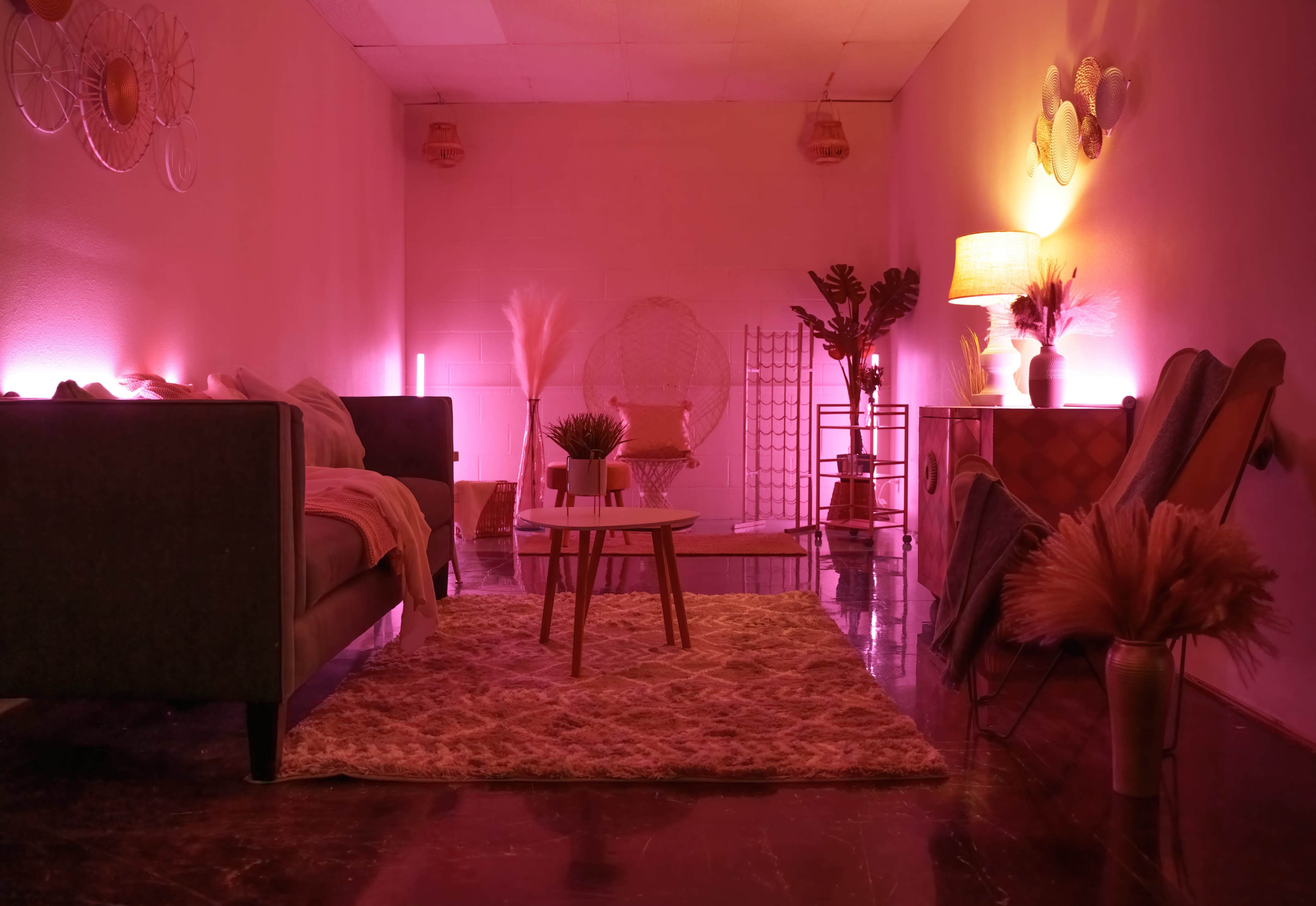 A softly lit room with a seating area featuring a dark sofa and chairs, a round table, decorative plants, and a rug.