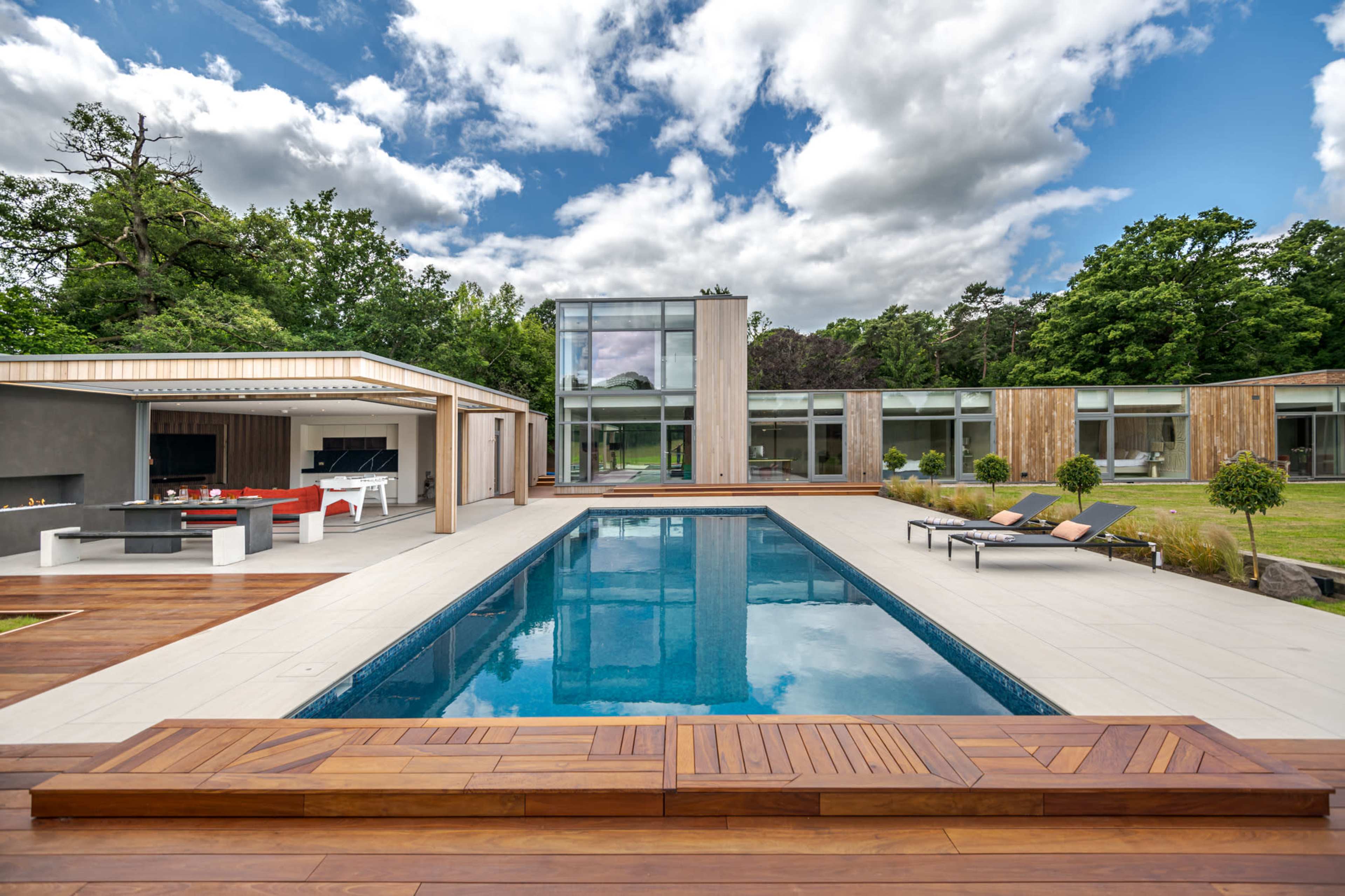 The image shows a modern house with large glass windows surrounding a swimming pool, complemented by a wooden deck, outdoor seating areas, and landscaped greenery.