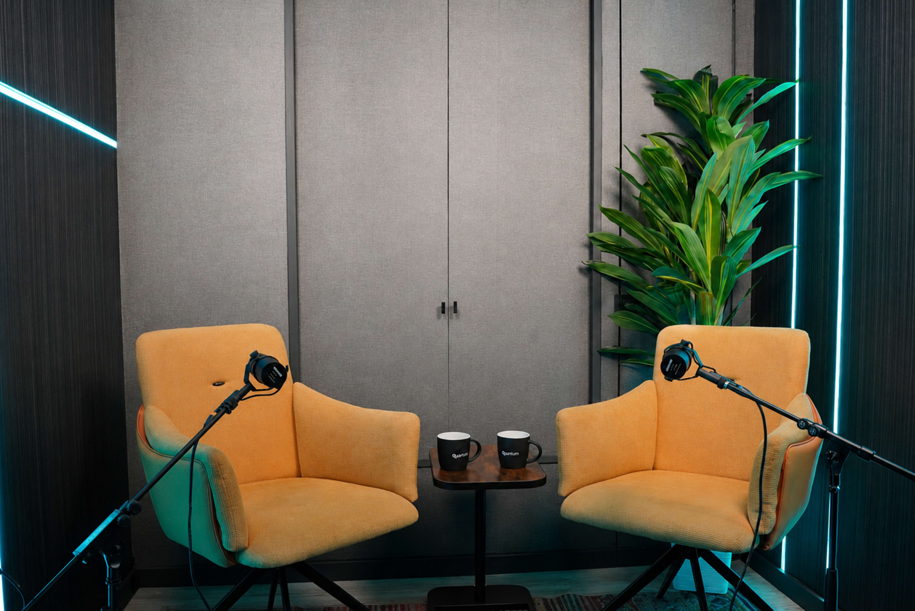 The image shows a well-lit podcast studio featuring two yellow chairs facing each other, with a small table between them, and a plant in the corner.