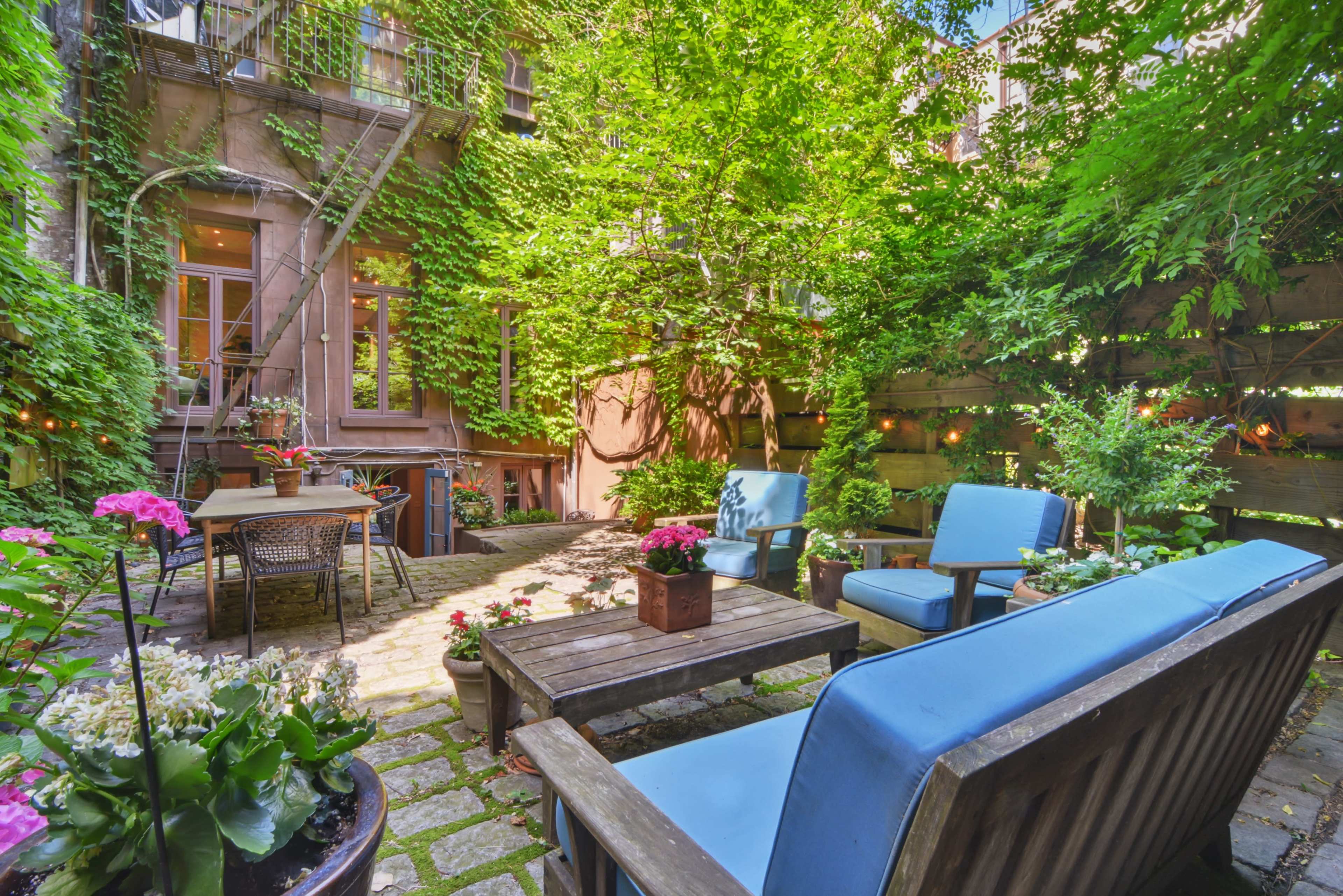 A small, landscaped courtyard features blue outdoor furniture, a wooden table, and potted plants, surrounded by ivy-covered walls.