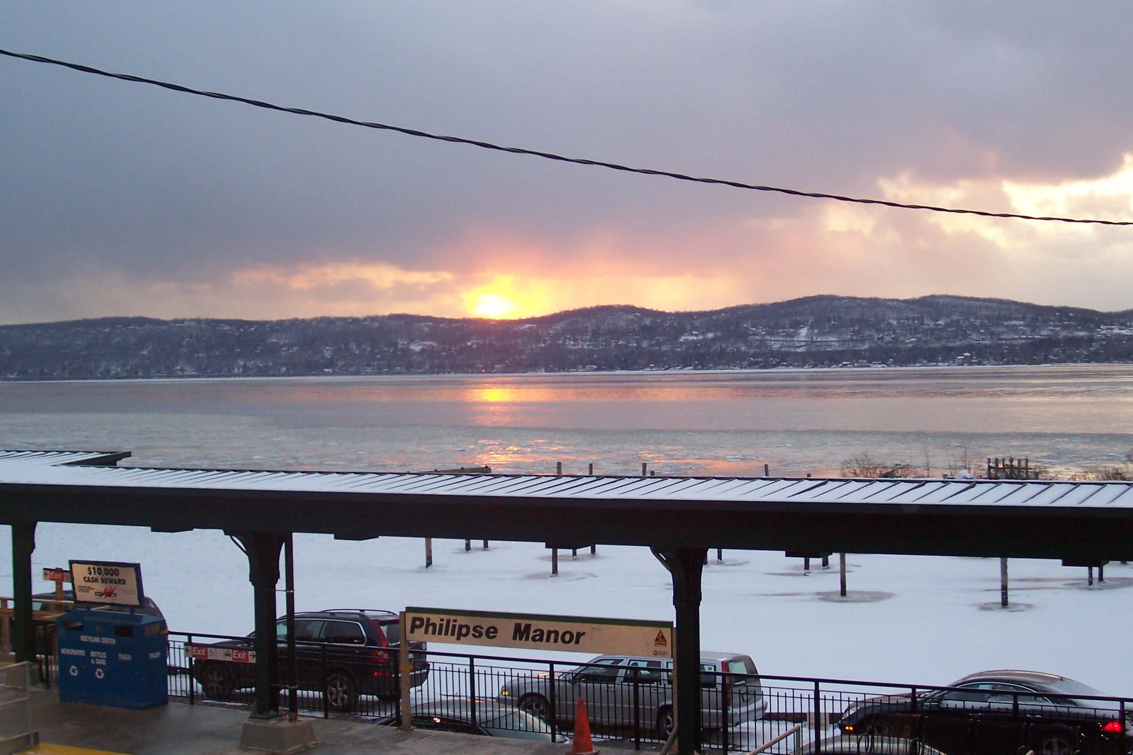The image shows a snow-covered shoreline by a river at sunset, with a train station sign reading "Philipse Manor" in the foreground.