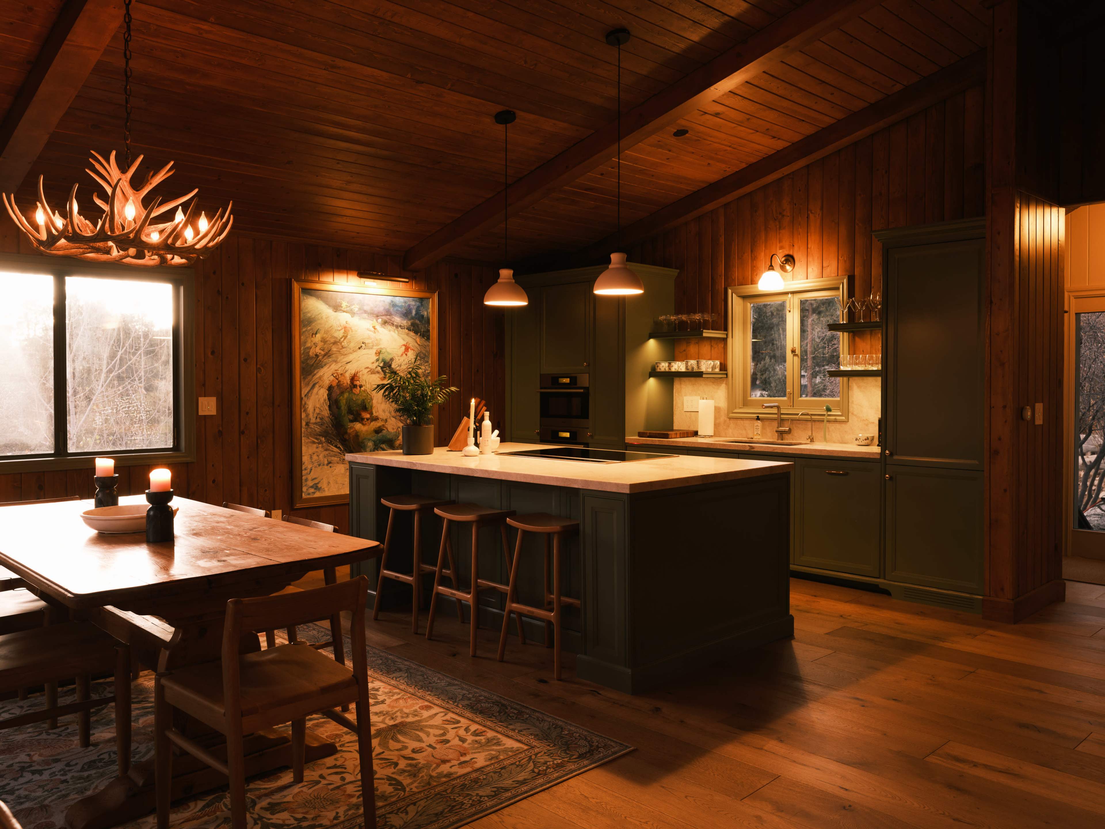 The image features a wooden kitchen with a central island, bar stools, and a dining table, all illuminated by warm lighting.