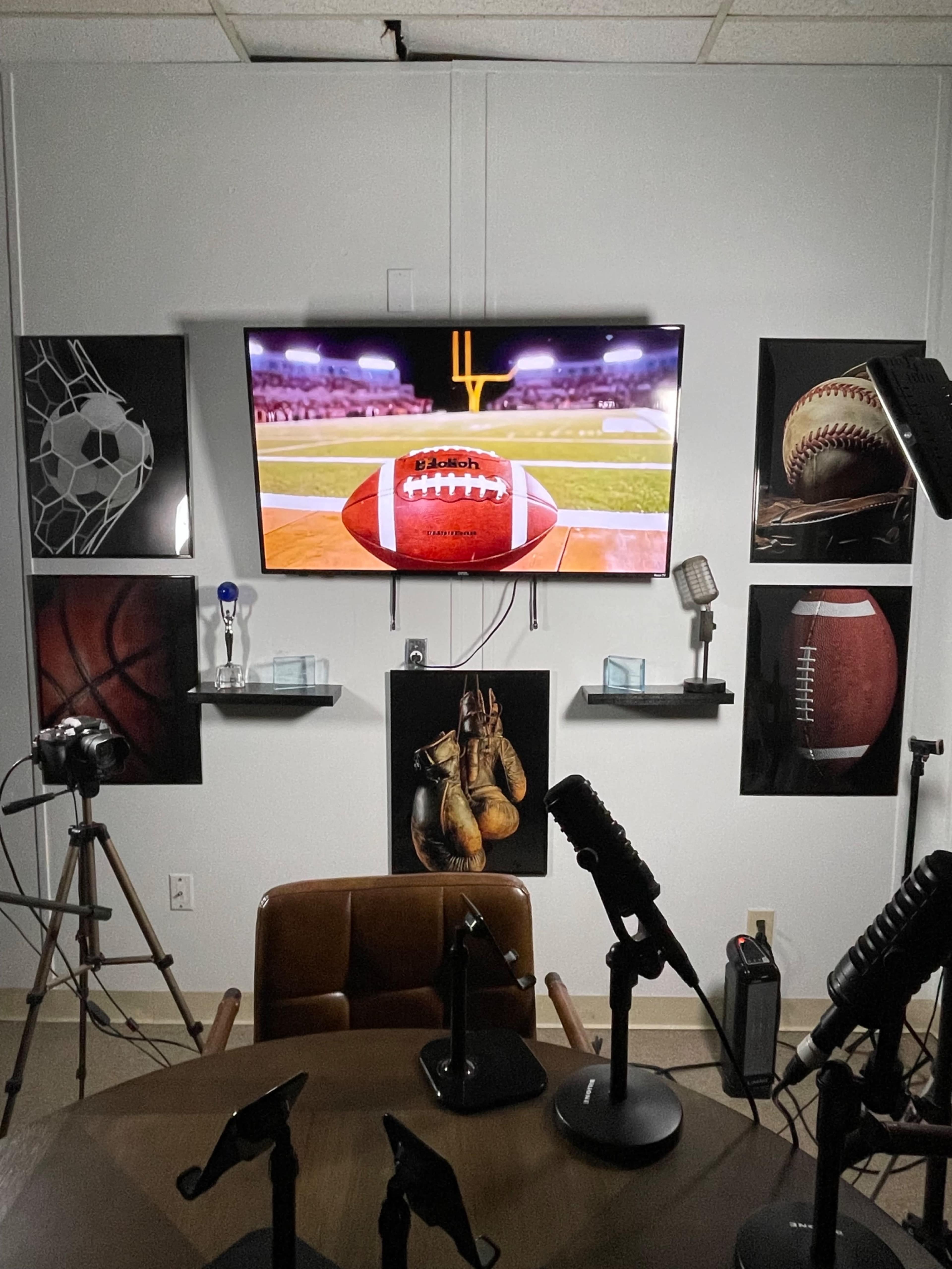 The image depicts a sports-themed studio featuring a wall-mounted television displaying a football game, surrounded by framed sports imagery, and a central table with microphones and cameras set up for recording.