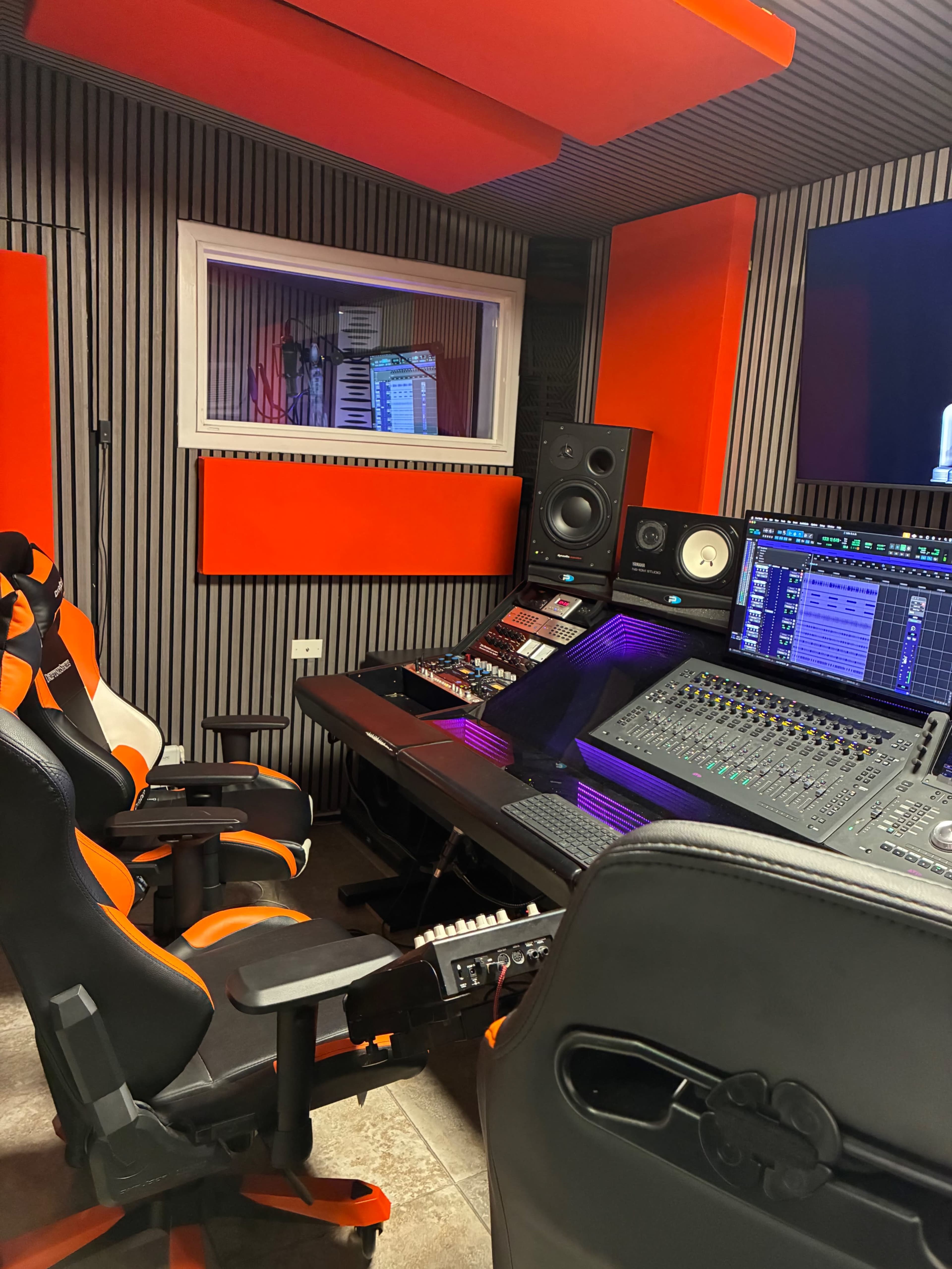 The image shows a music production studio with a soundboard, two gaming-style chairs, and soundproofing panels on the walls.