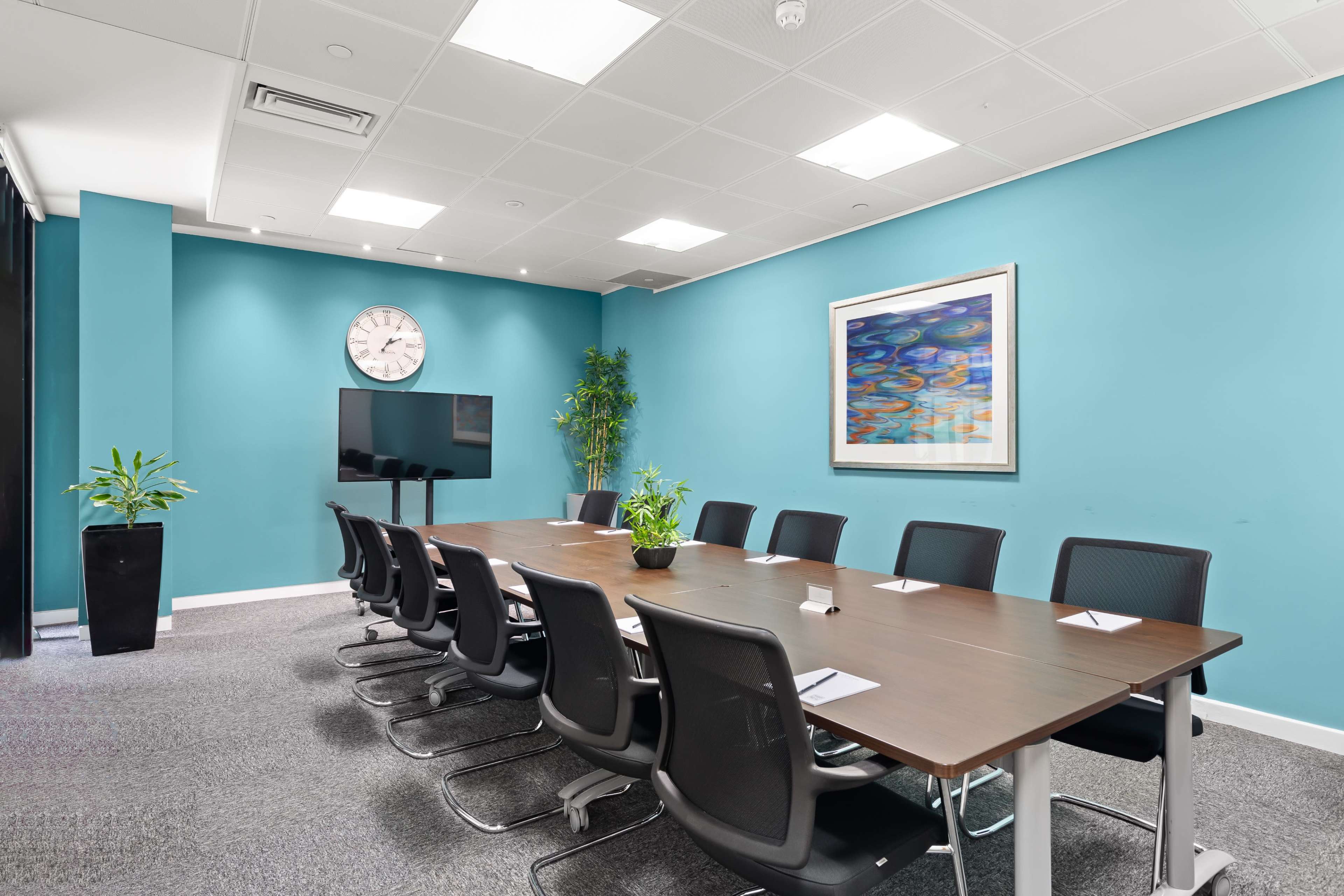 A modern conference room features a long wooden table surrounded by black chairs, a large television, a wall clock, and colorful artwork, all set against a vibrant blue wall.