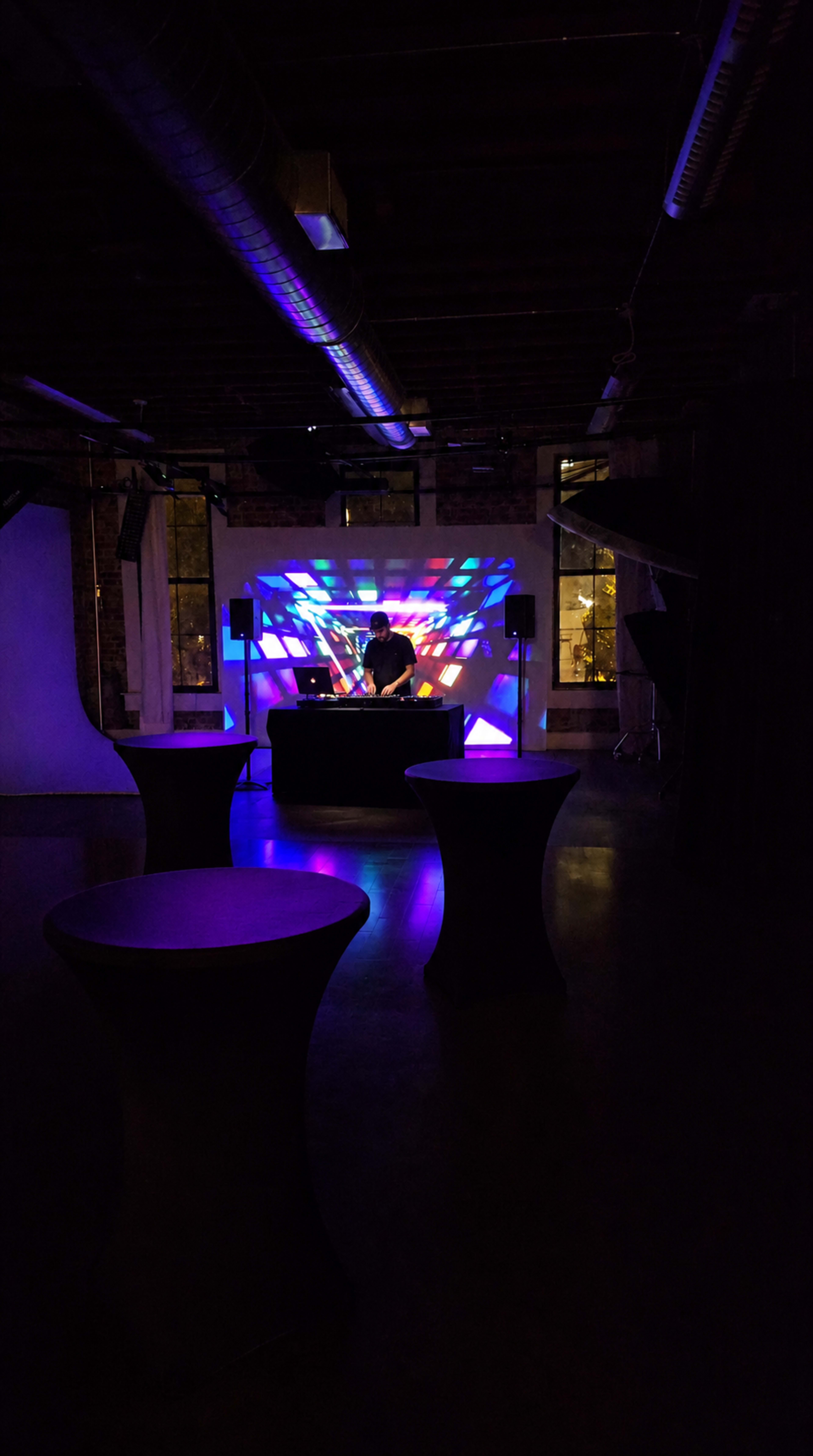 A DJ stands behind a table while colorful lights project patterns onto a wall in a dimly lit indoor space.