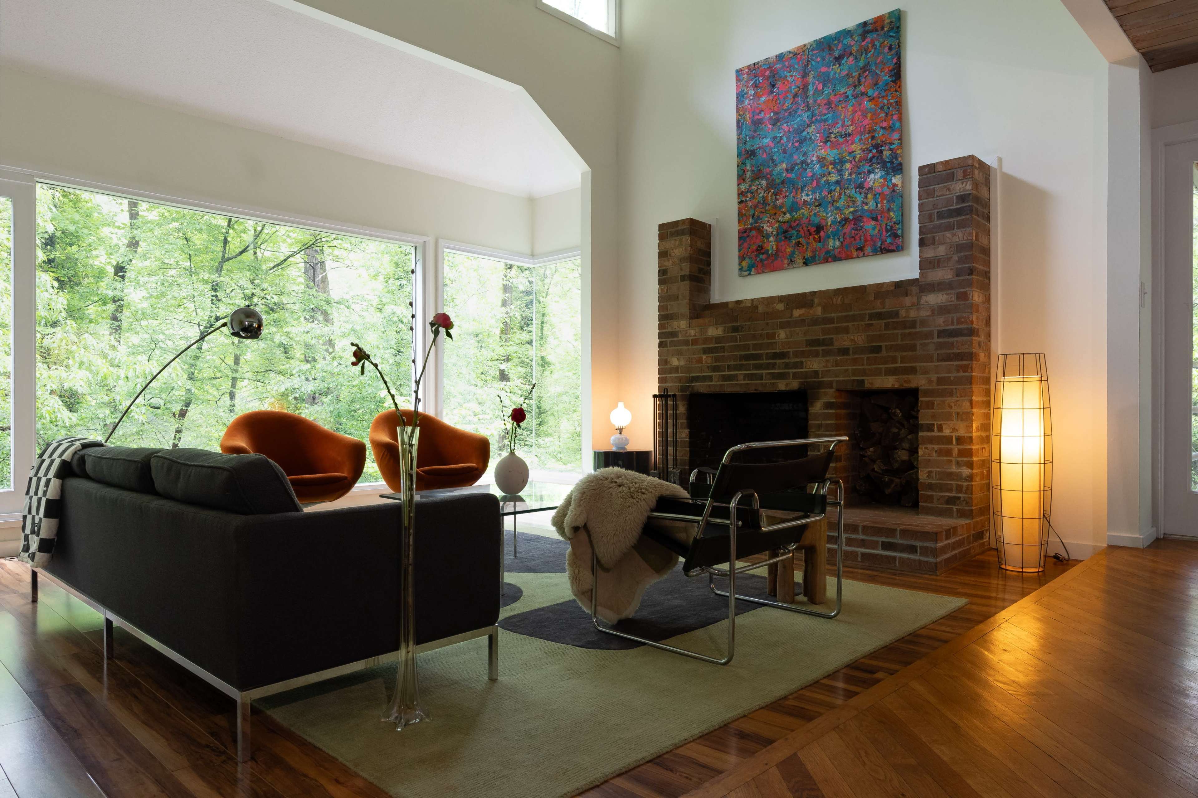 A modern living room features large windows with a view of greenery, two orange chairs, a black couch, and a brick fireplace with a colorful artwork above it.