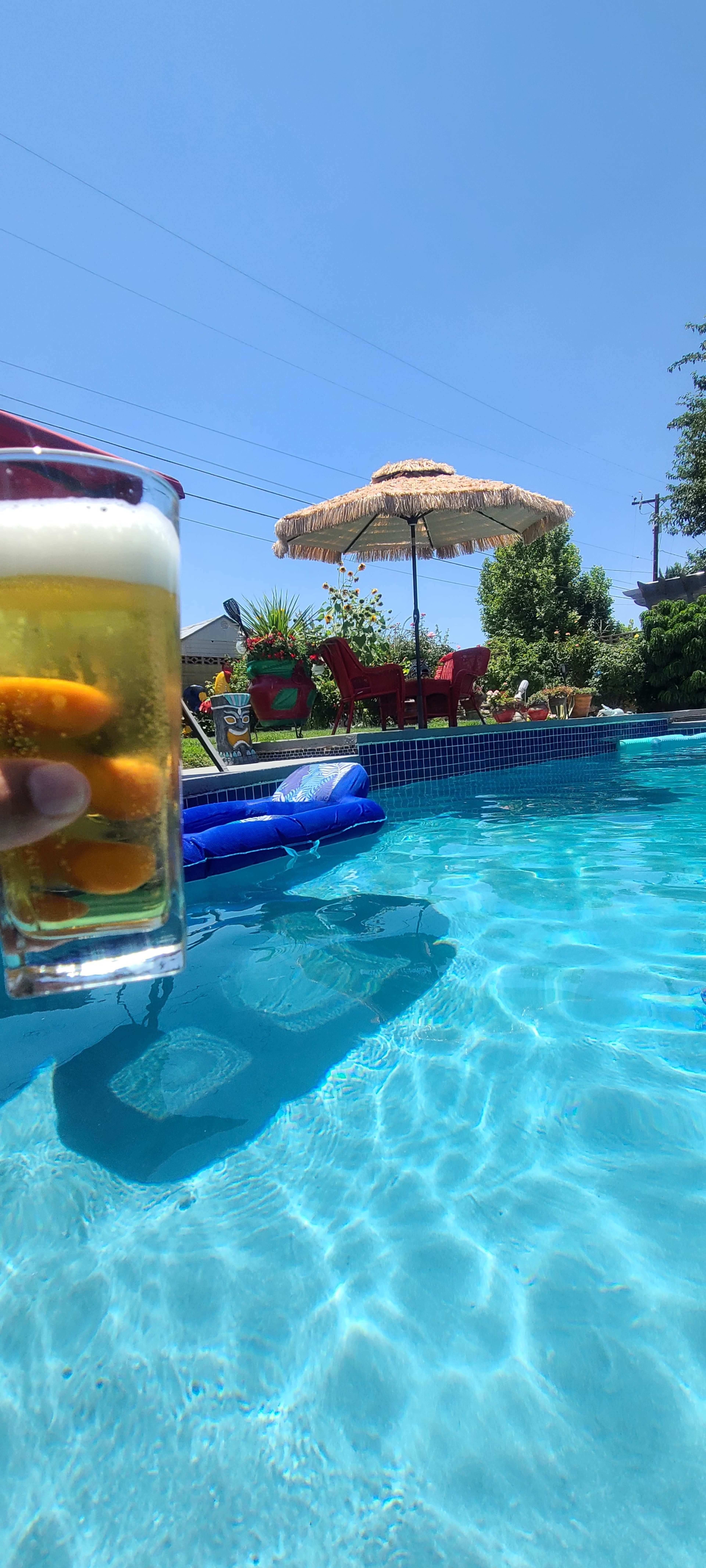 A hand holds a glass of beer above a clear swimming pool with a floating lounge and a shaded seating area in the background.