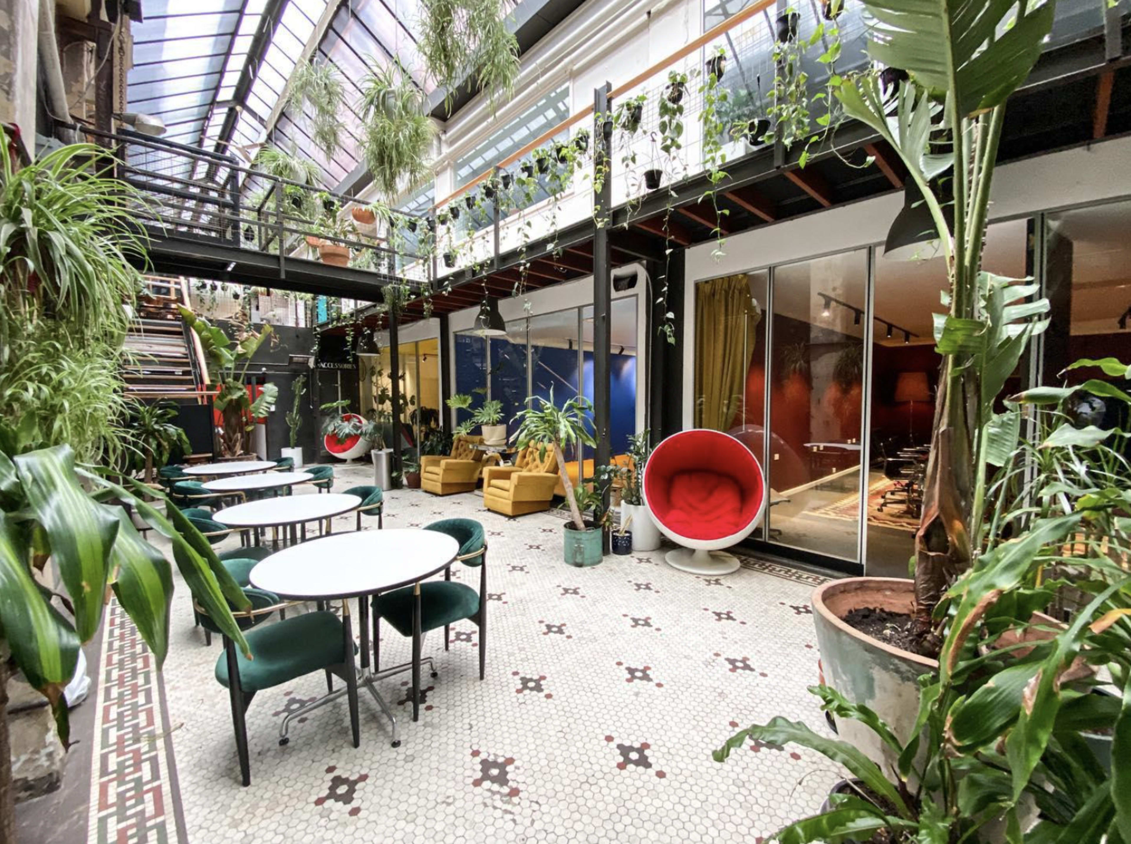 A modern indoor space filled with various plants, featuring green chairs and tables, large windows, and vibrant decor.