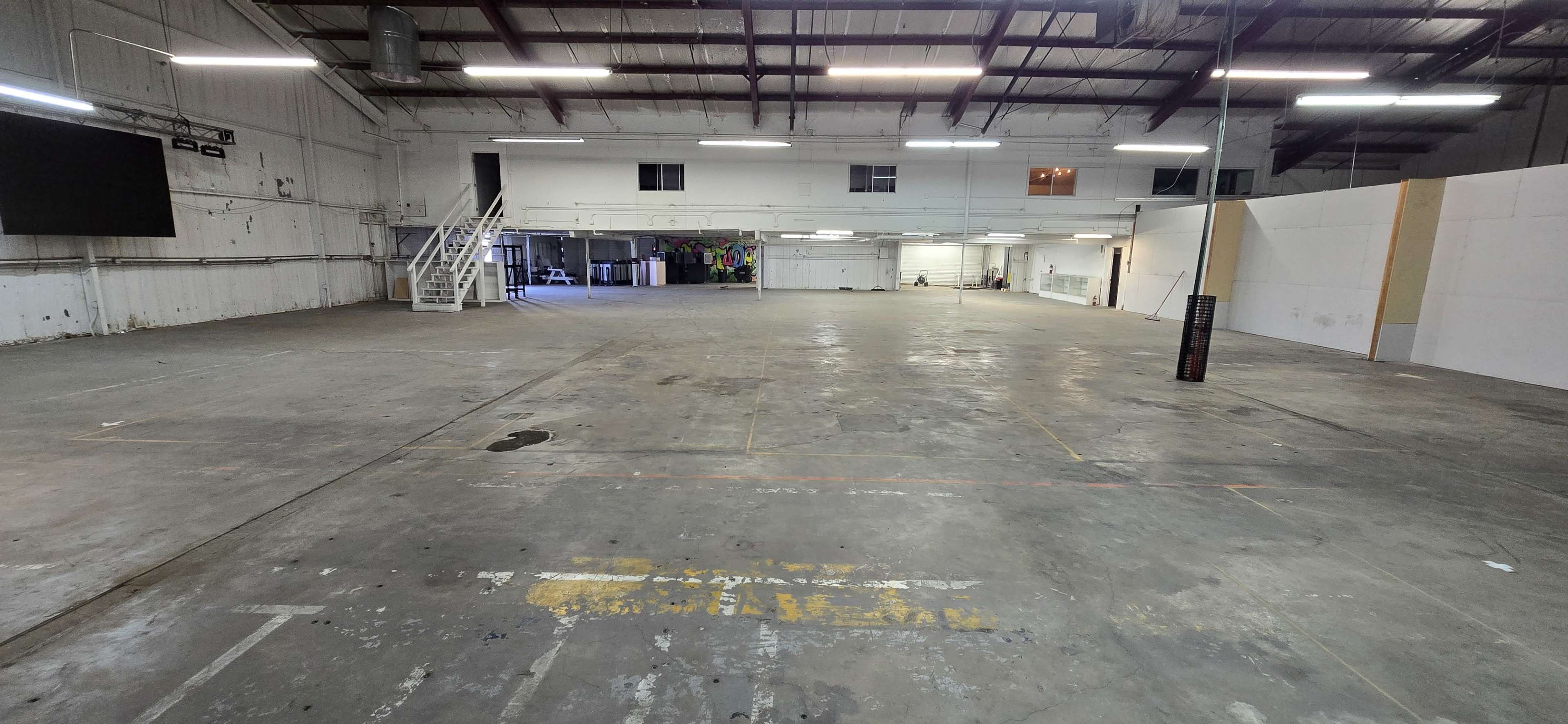 The image shows a large, empty industrial warehouse space with concrete flooring and minimal furnishings.