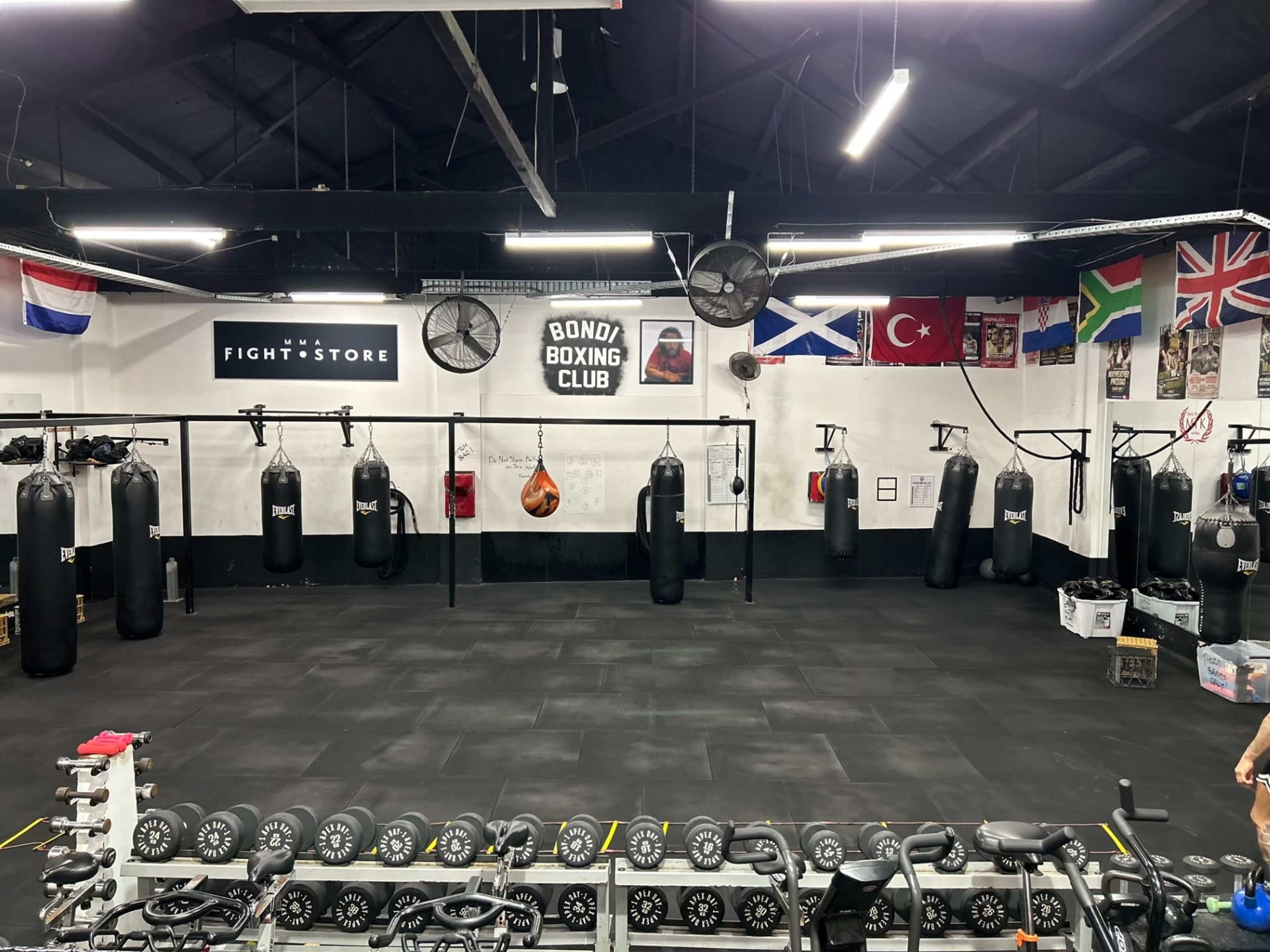 An empty boxing gym with hanging punching bags, a punching bag on the floor, and a row of dumbbells in the foreground.
