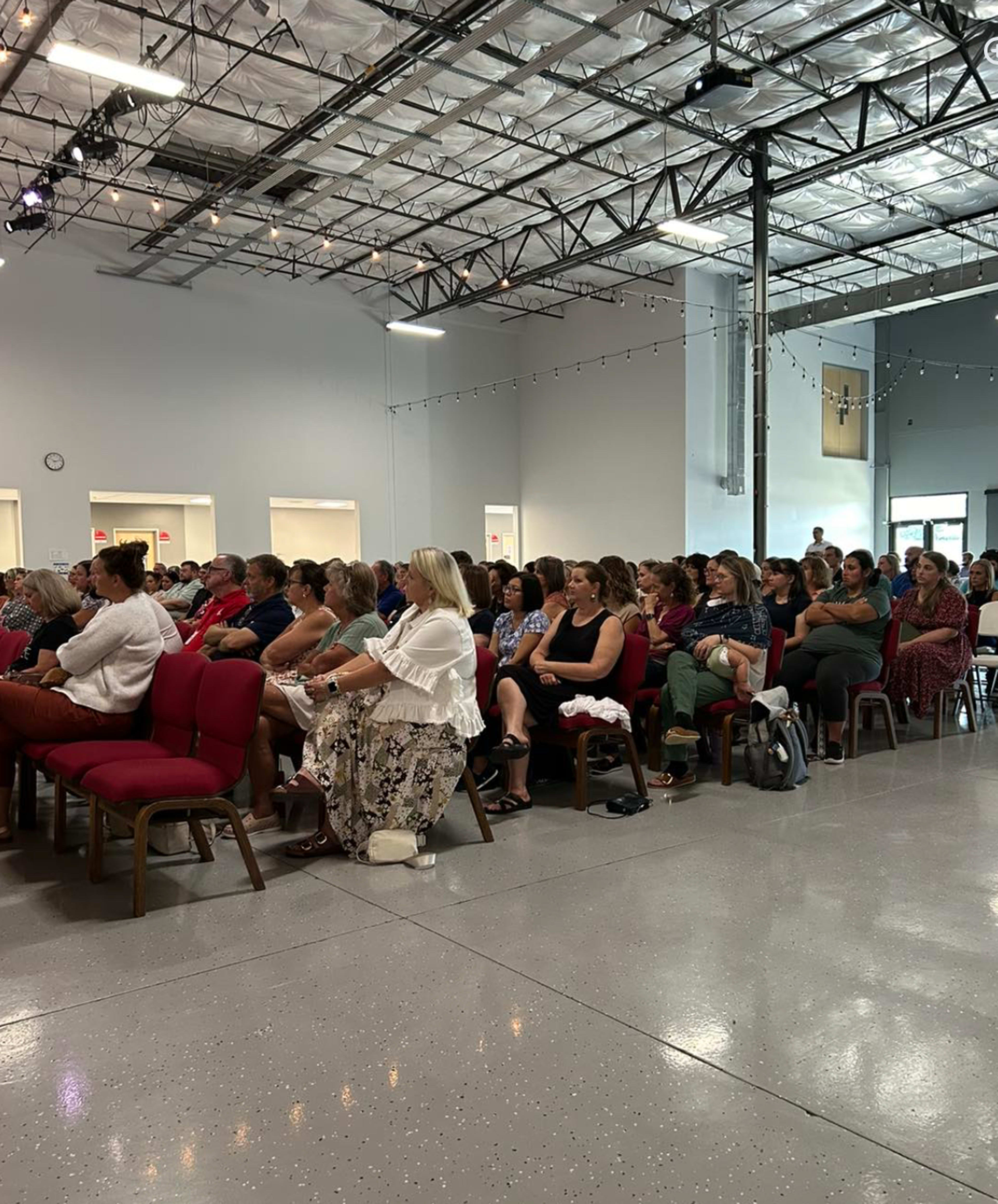 A large audience is seated in a well-lit indoor space, attentively facing a stage area.