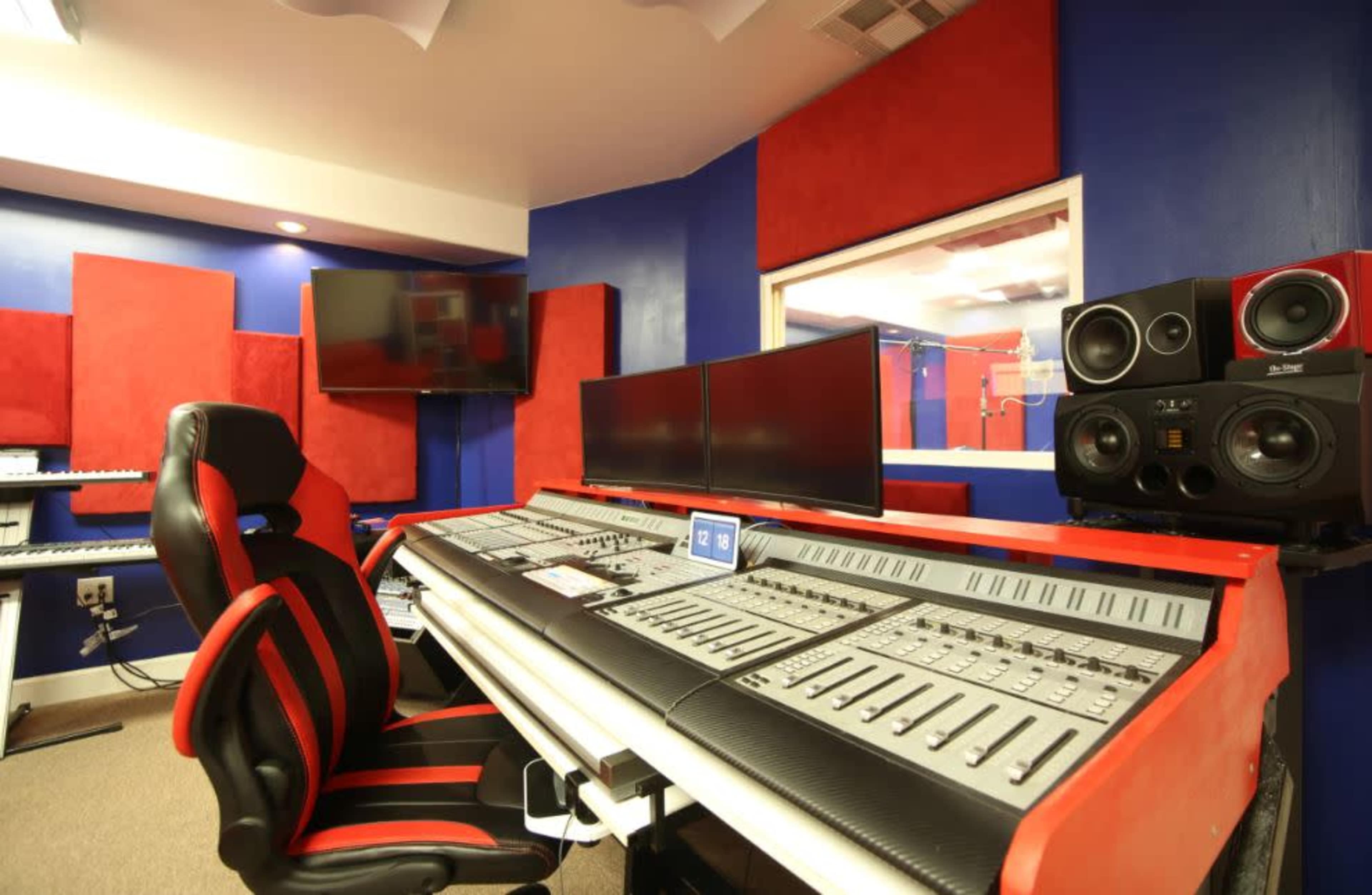 The image shows a music production studio with a large mixing console, black and red acoustic panels, multiple monitors, and a comfortable black and red office chair.