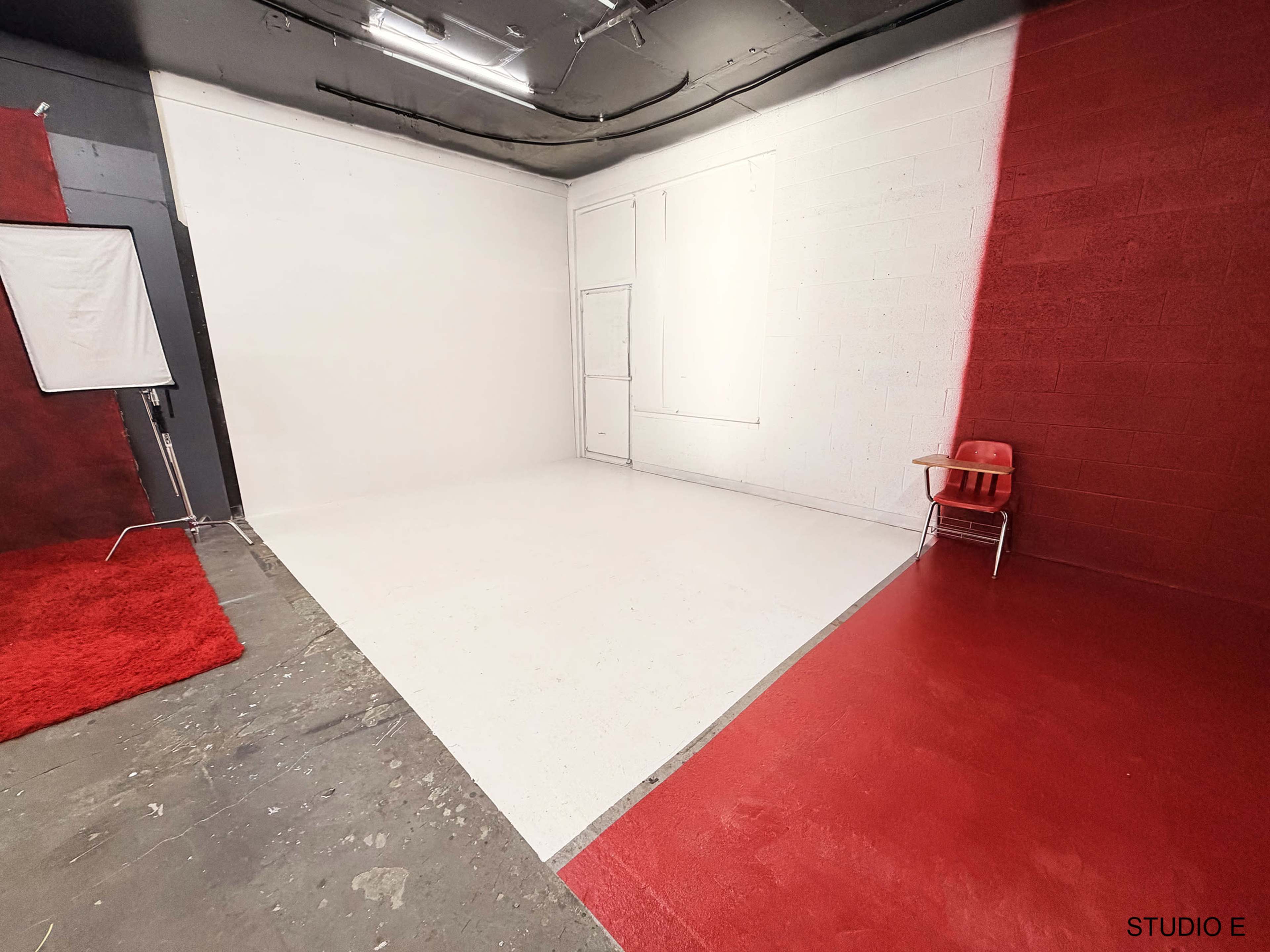 The image shows a studio space with a white wall on one side and a red wall on the other, featuring a red chair and a white floor.