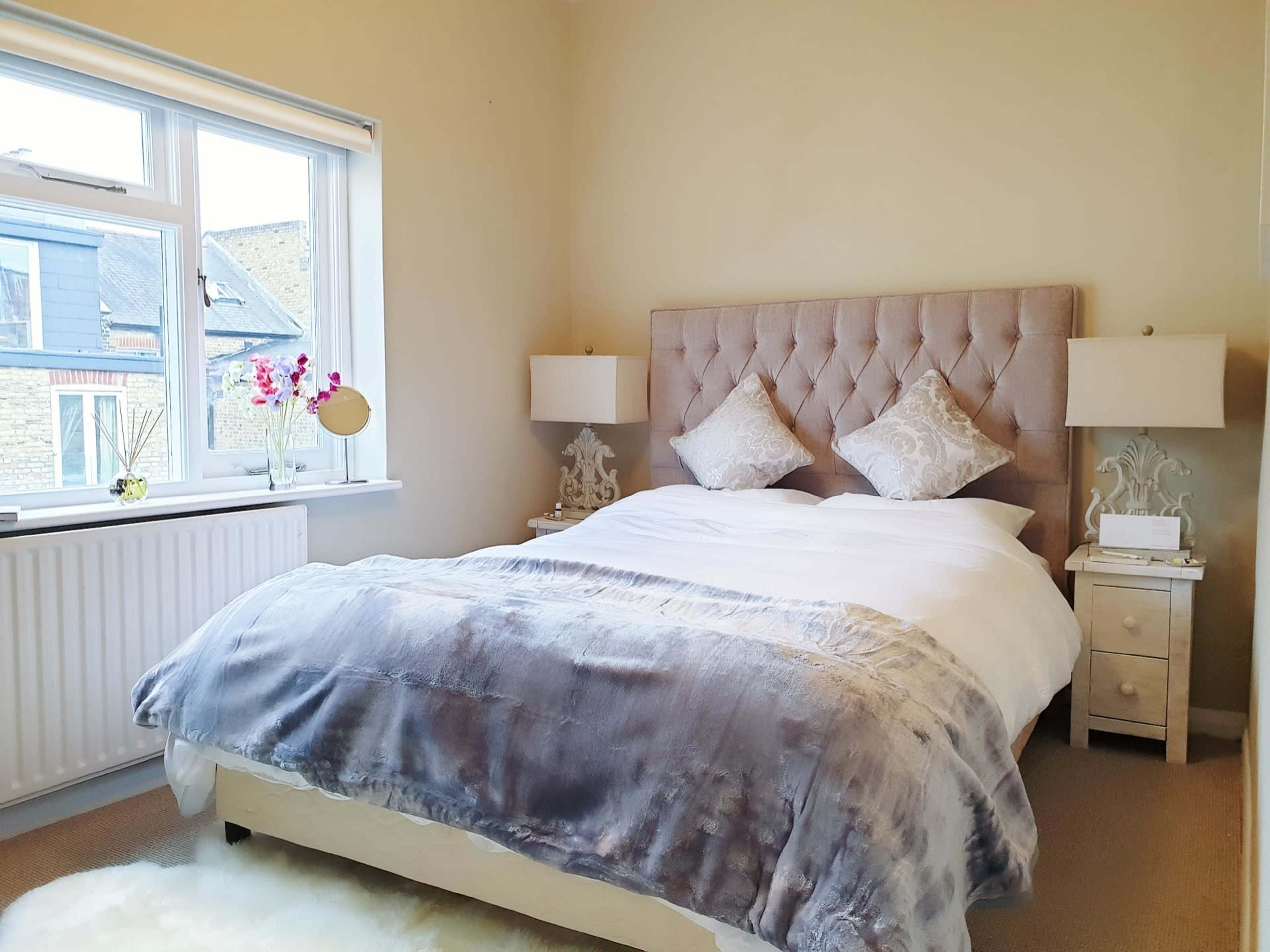 A neatly arranged bedroom features a large bed with a tufted headboard, two bedside tables with lamps, and a window allowing natural light.