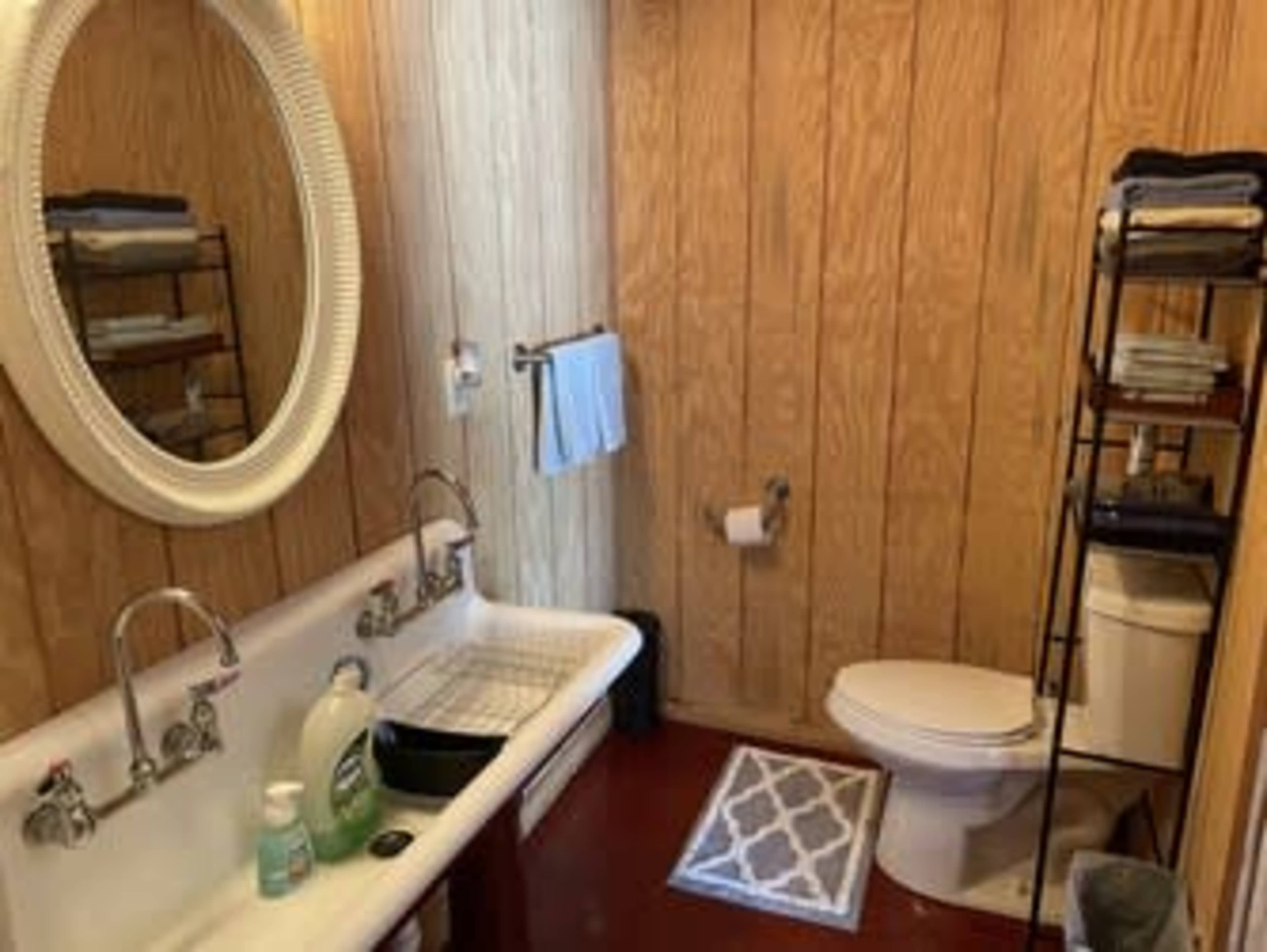 The bathroom features a vintage sink with a countertop, a toilet, a shelf with neatly arranged towels, and wooden paneling on the walls.