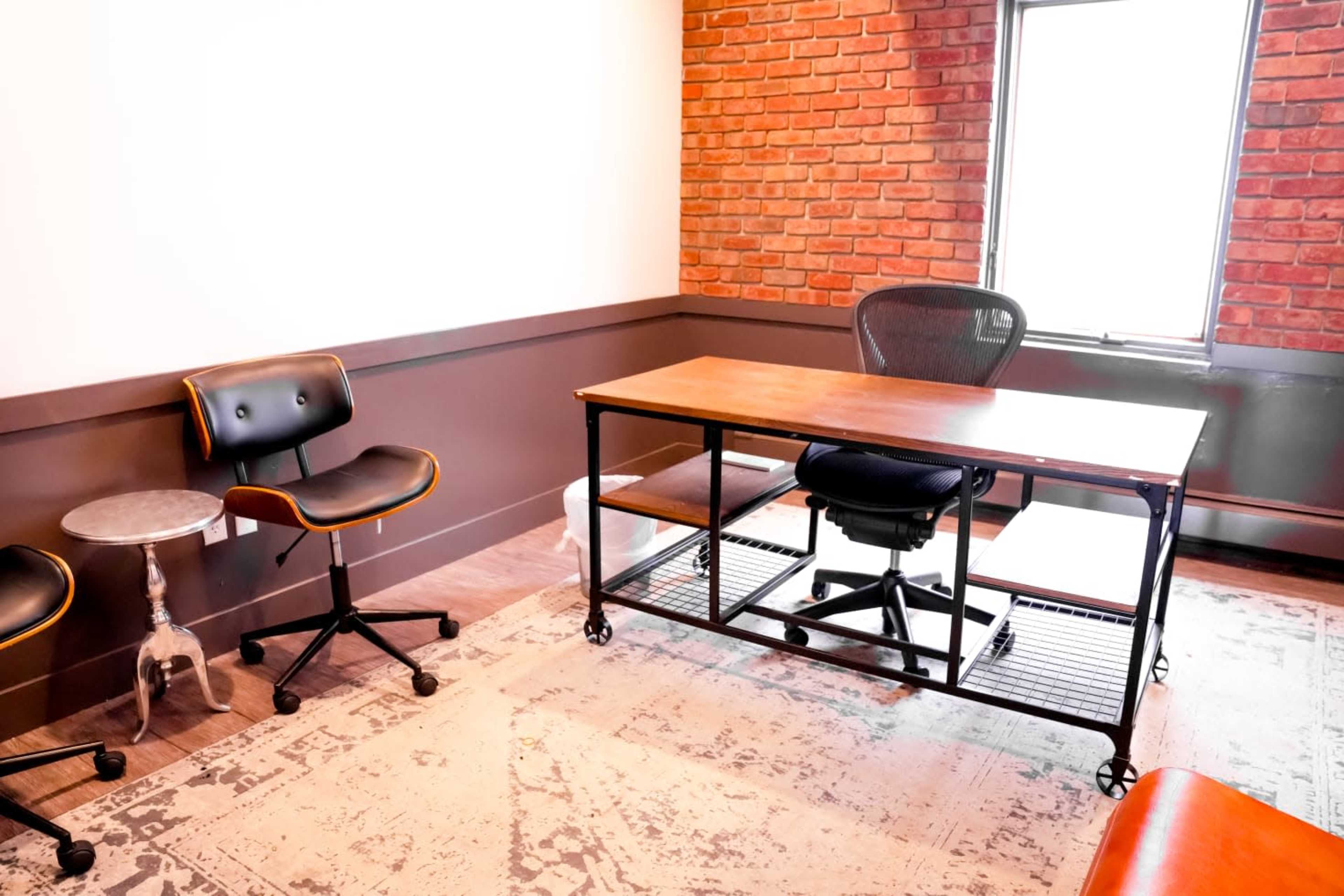 A workspace featuring a wooden desk on wheels with a black office chair, two black rolling chairs, and a small side table in a room with a brick accent wall.