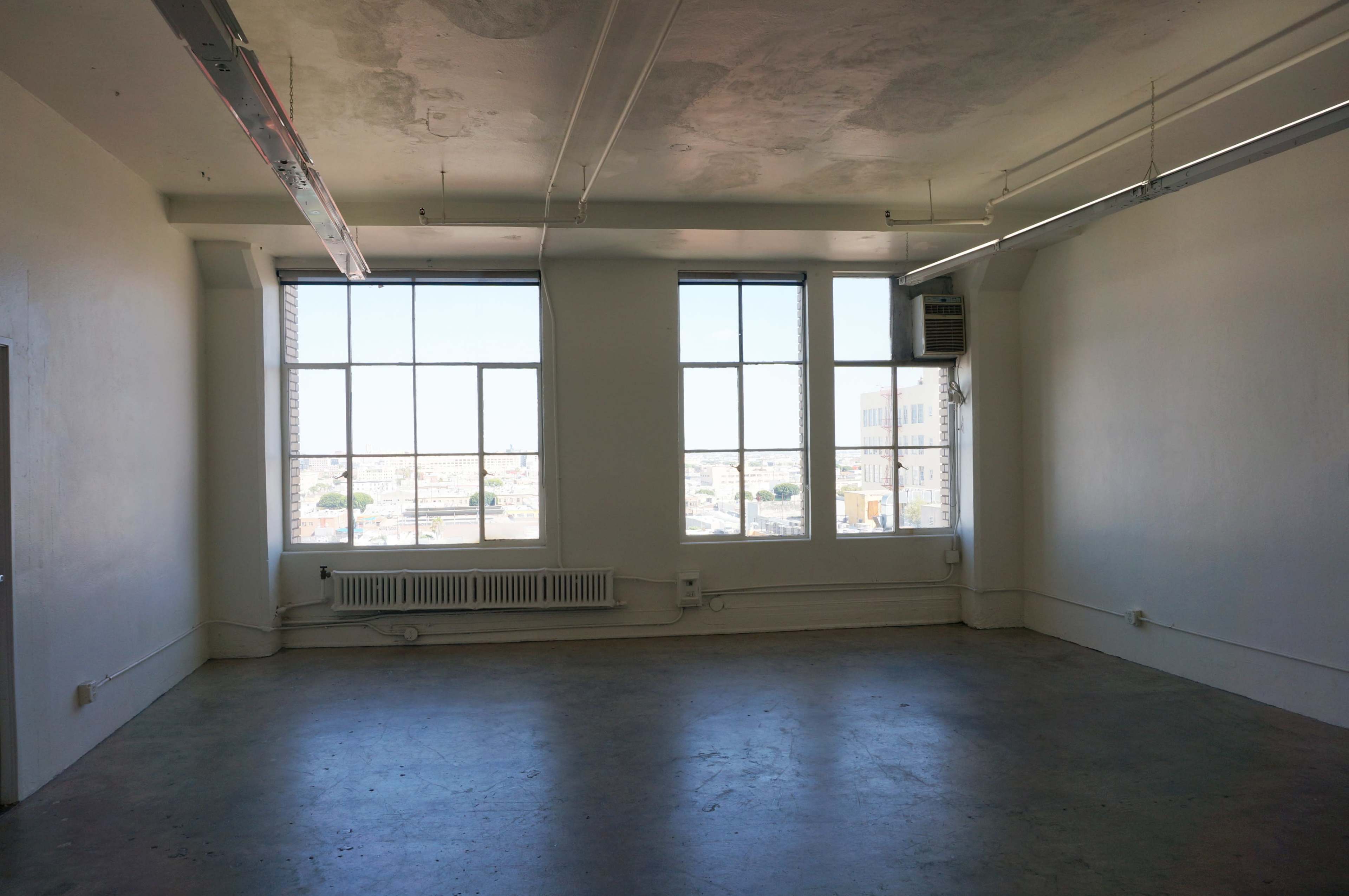 The image shows an empty room with large windows allowing natural light to enter.