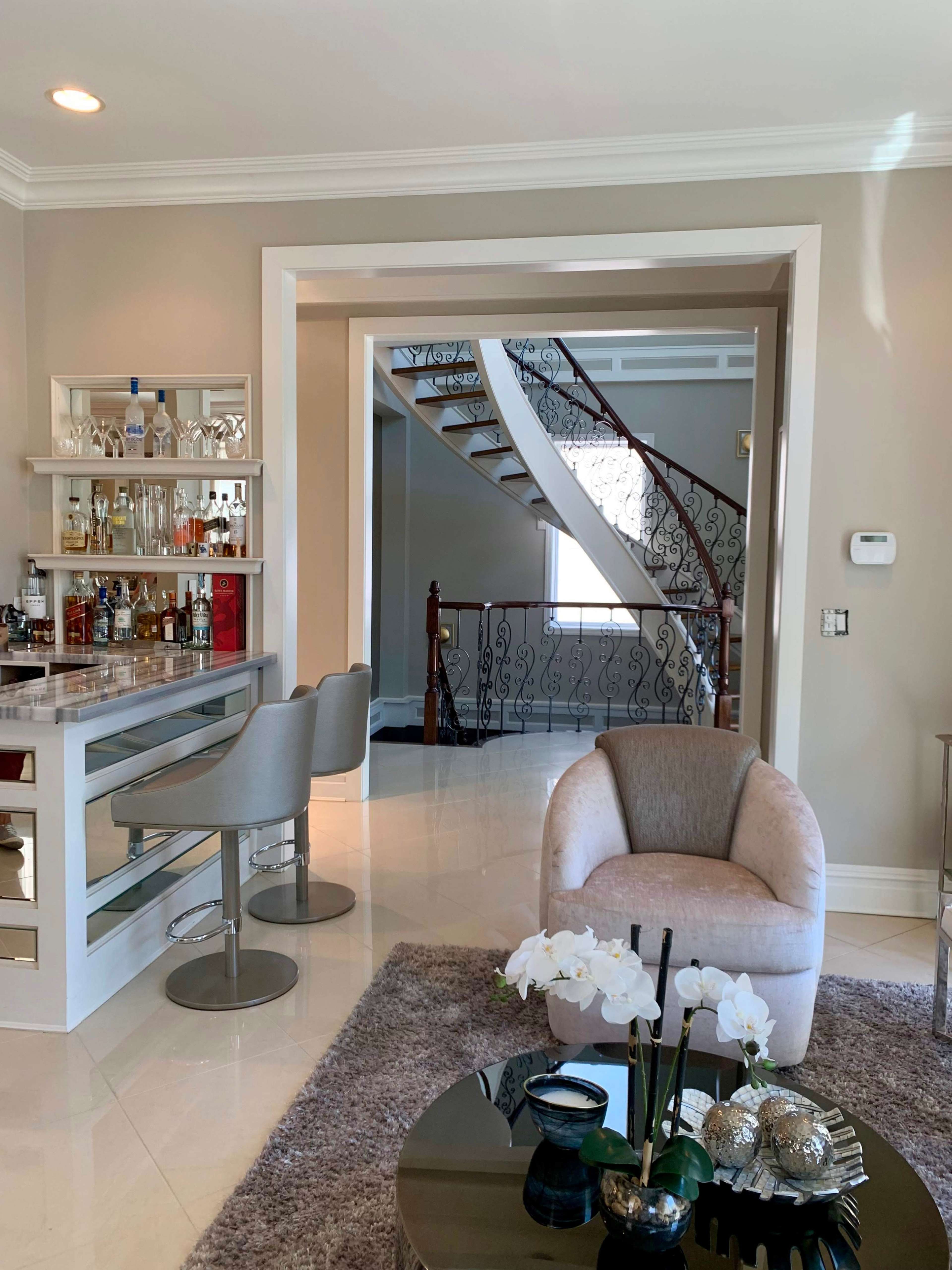 A modern interior with a home bar area, a round table with decorative flowers, and a staircase in the background.