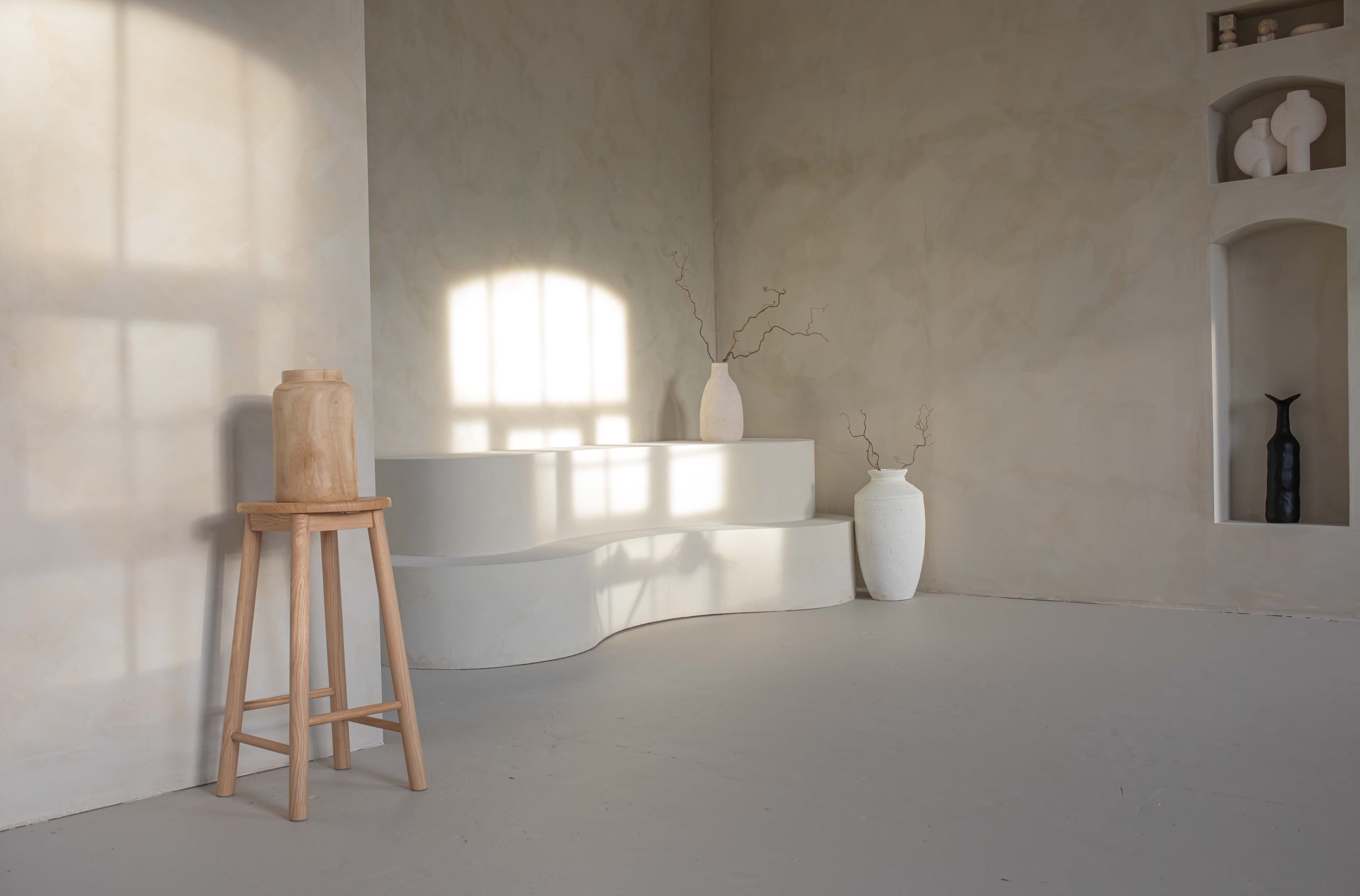 The image shows a minimalist interior space featuring a curved white platform, a wooden stool, and several vases placed against a softly textured beige wall.
