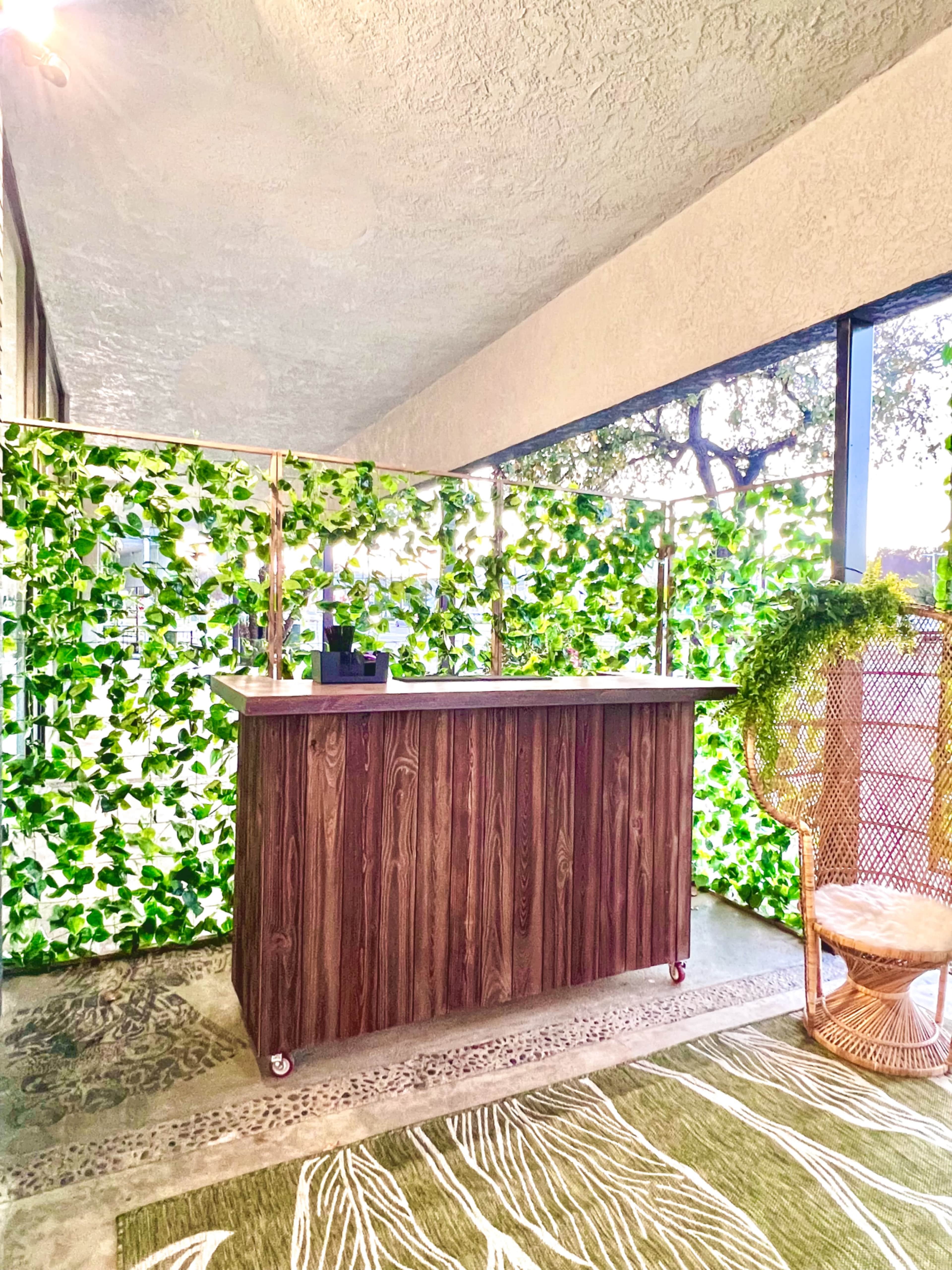 The scene features a wooden bar against a backdrop of lush green leaves, with a wicker chair and a patterned rug nearby.