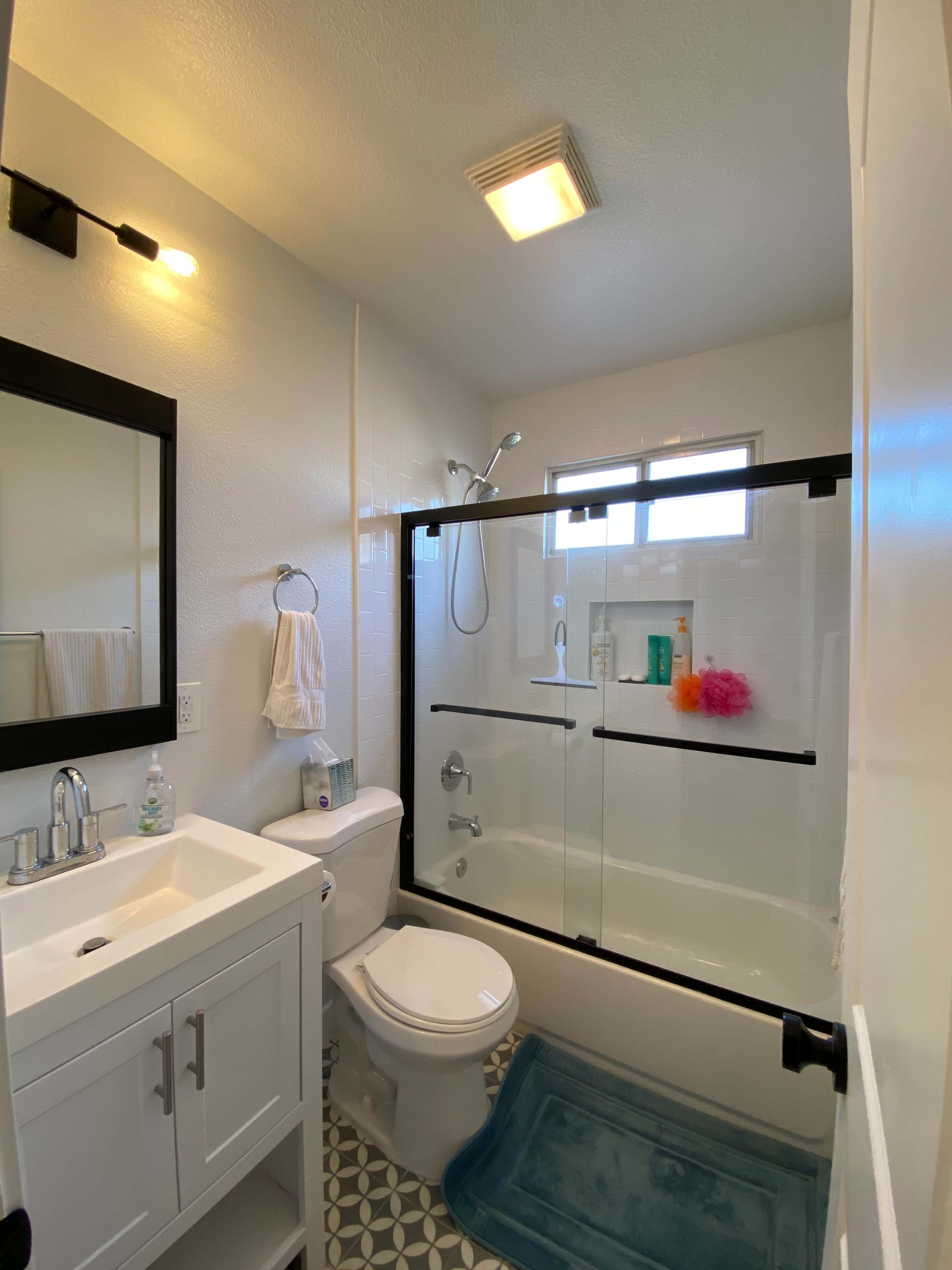 A small bathroom features a bathtub with a glass shower door, a white vanity, and a mirror, with patterned tile on the floor.