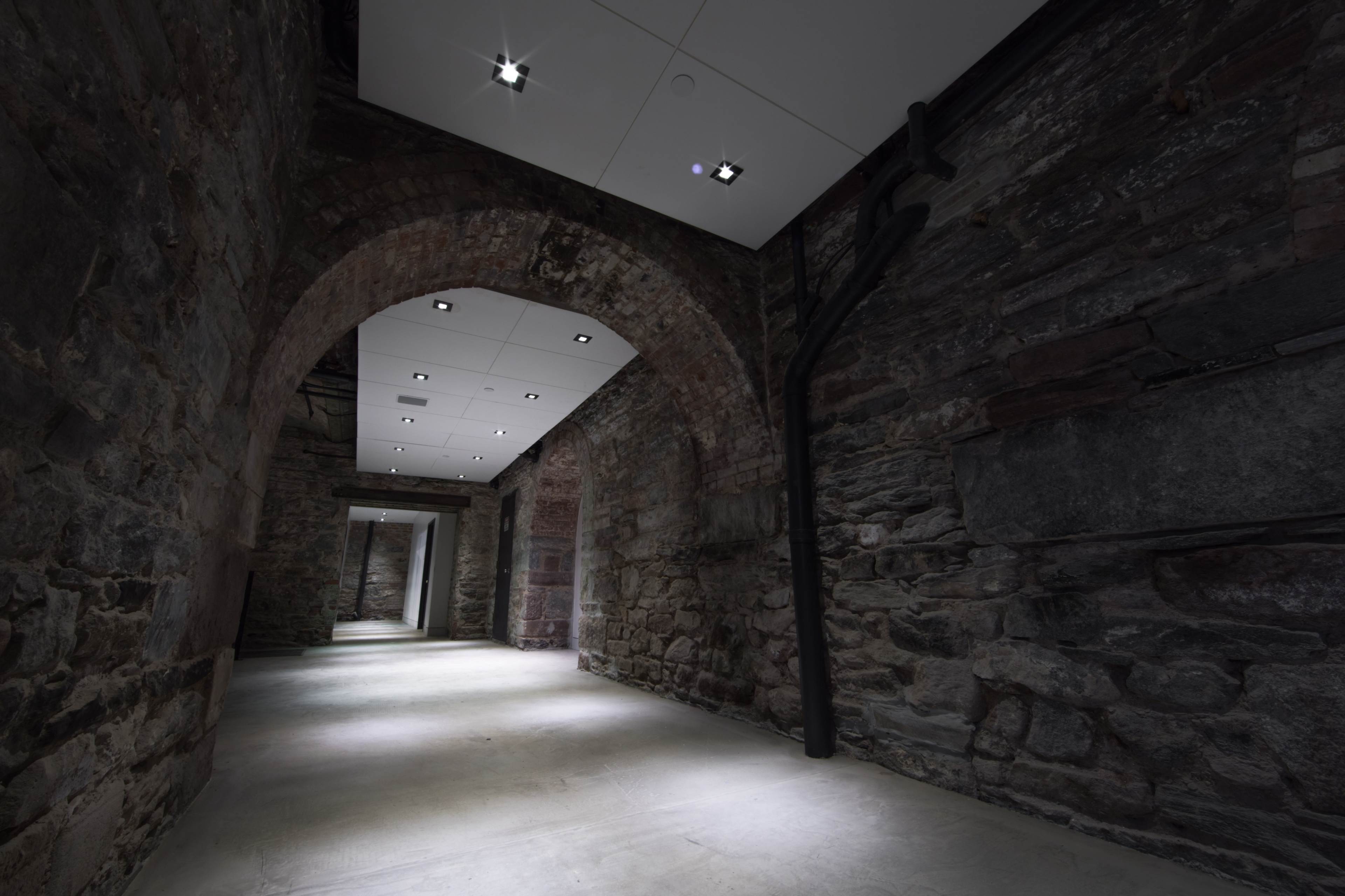 The image shows a dimly lit corridor with exposed stone walls and a ceiling that features modern lighting fixtures.