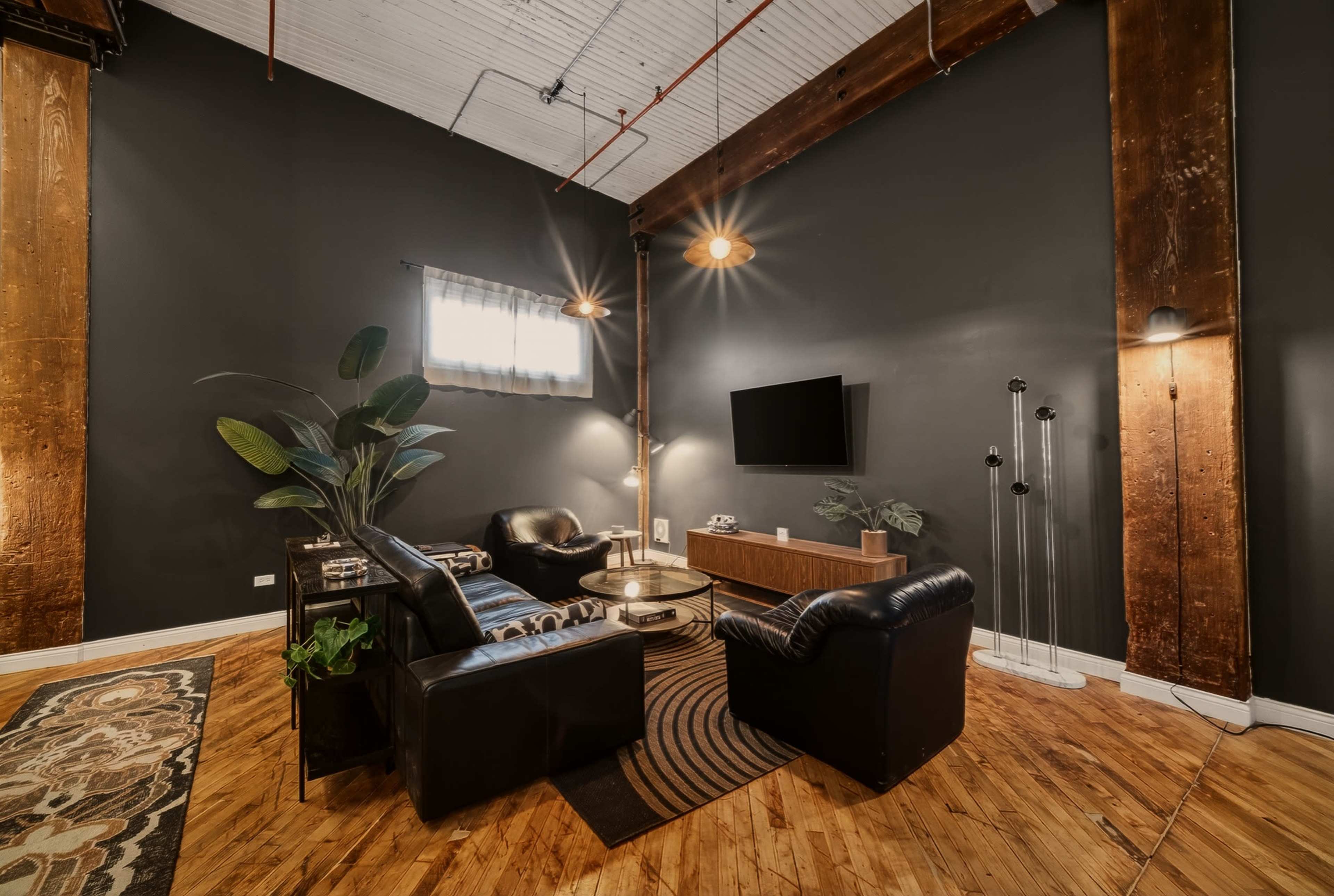 The image shows a modern sitting area with a black leather sofa set, a wooden coffee table, and a wall-mounted television, framed by exposed wooden beams and industrial lighting.