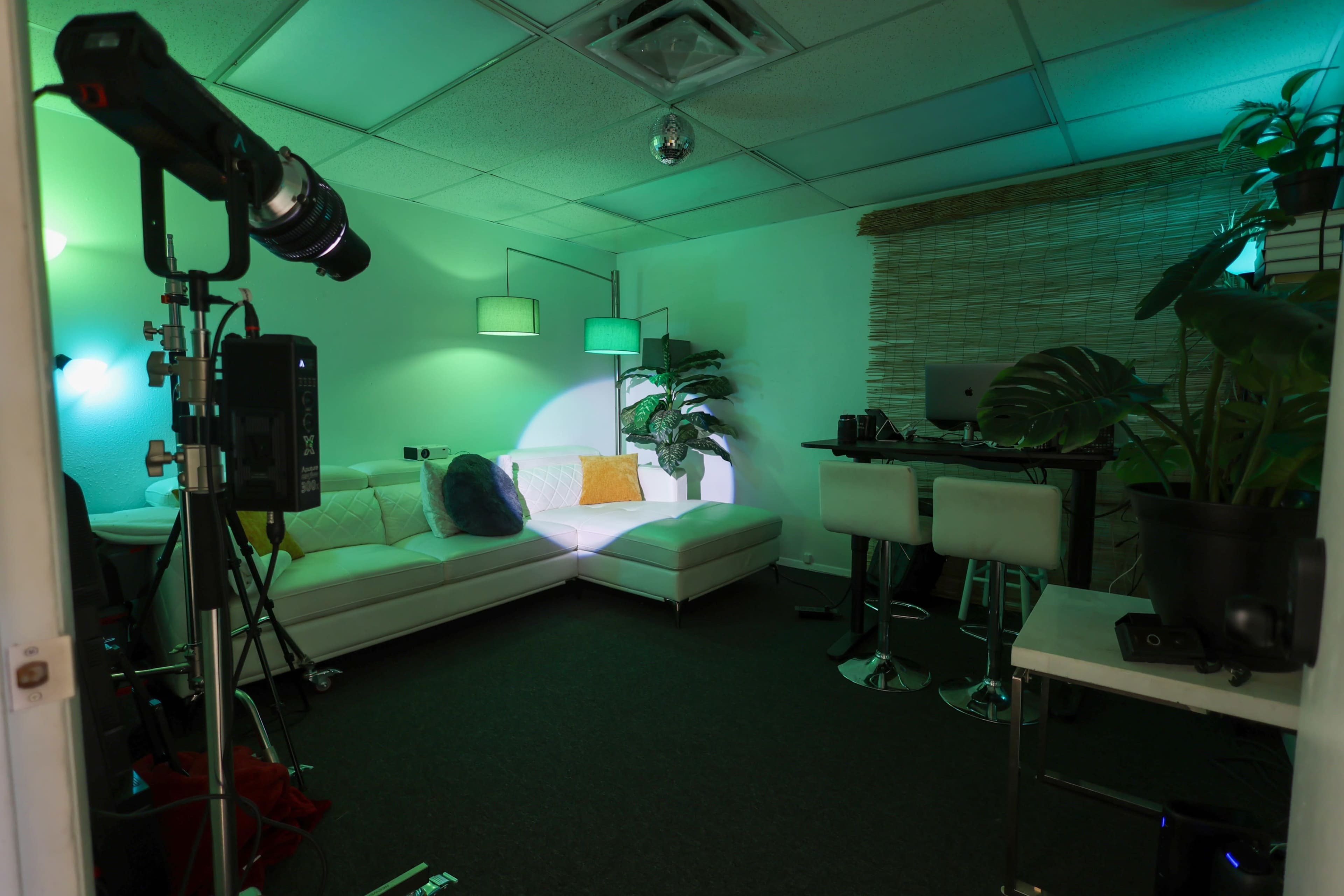 The image shows a modern lounge area with a white sectional sofa, bar stools, a small table, and decorative plants, all illuminated by green and white lighting.