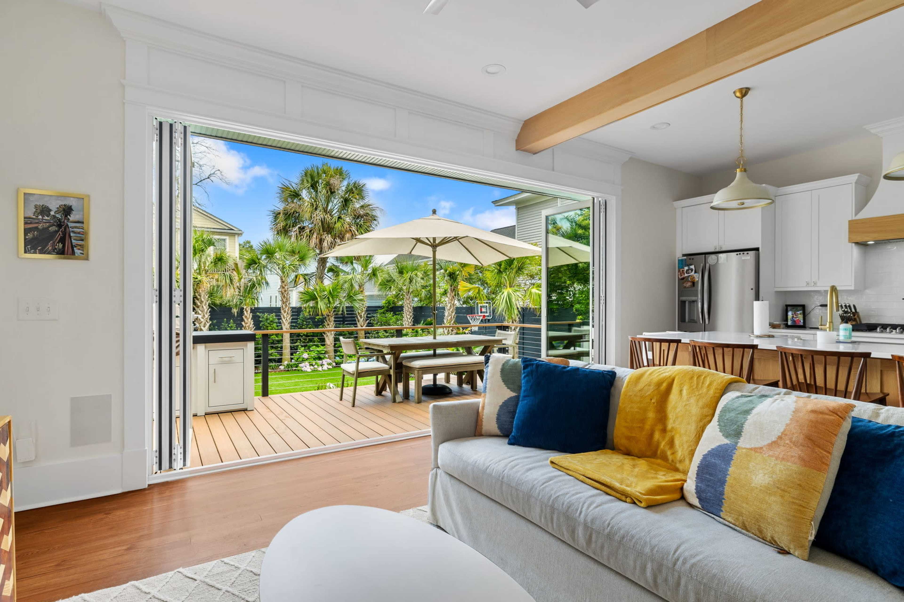 A living room features a sofa with colorful cushions, open sliding doors leading to a deck with outdoor seating and an umbrella, surrounded by palm trees.