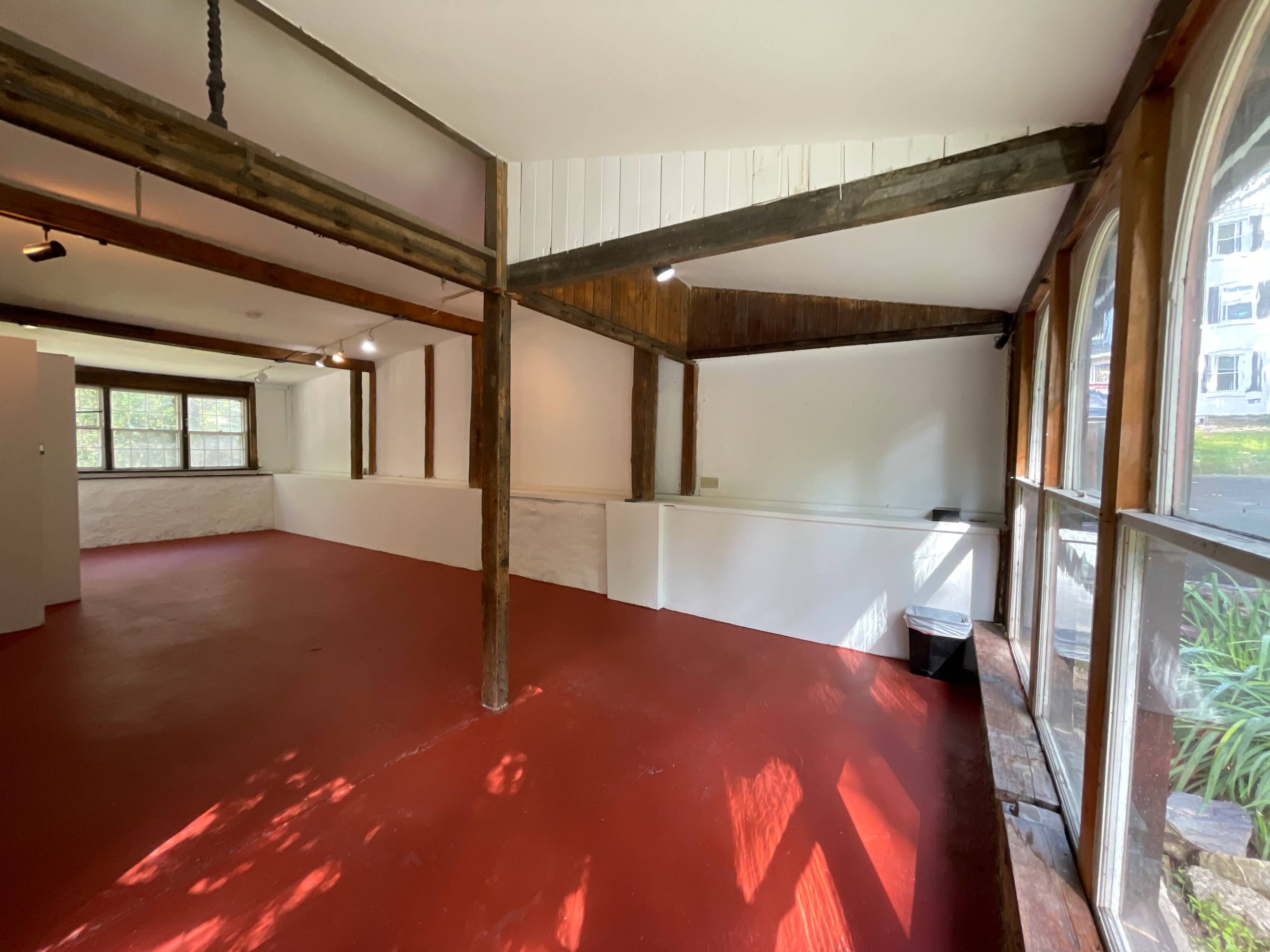 An interior space with red flooring and wooden beams, featuring white walls and large windows that allow natural light.