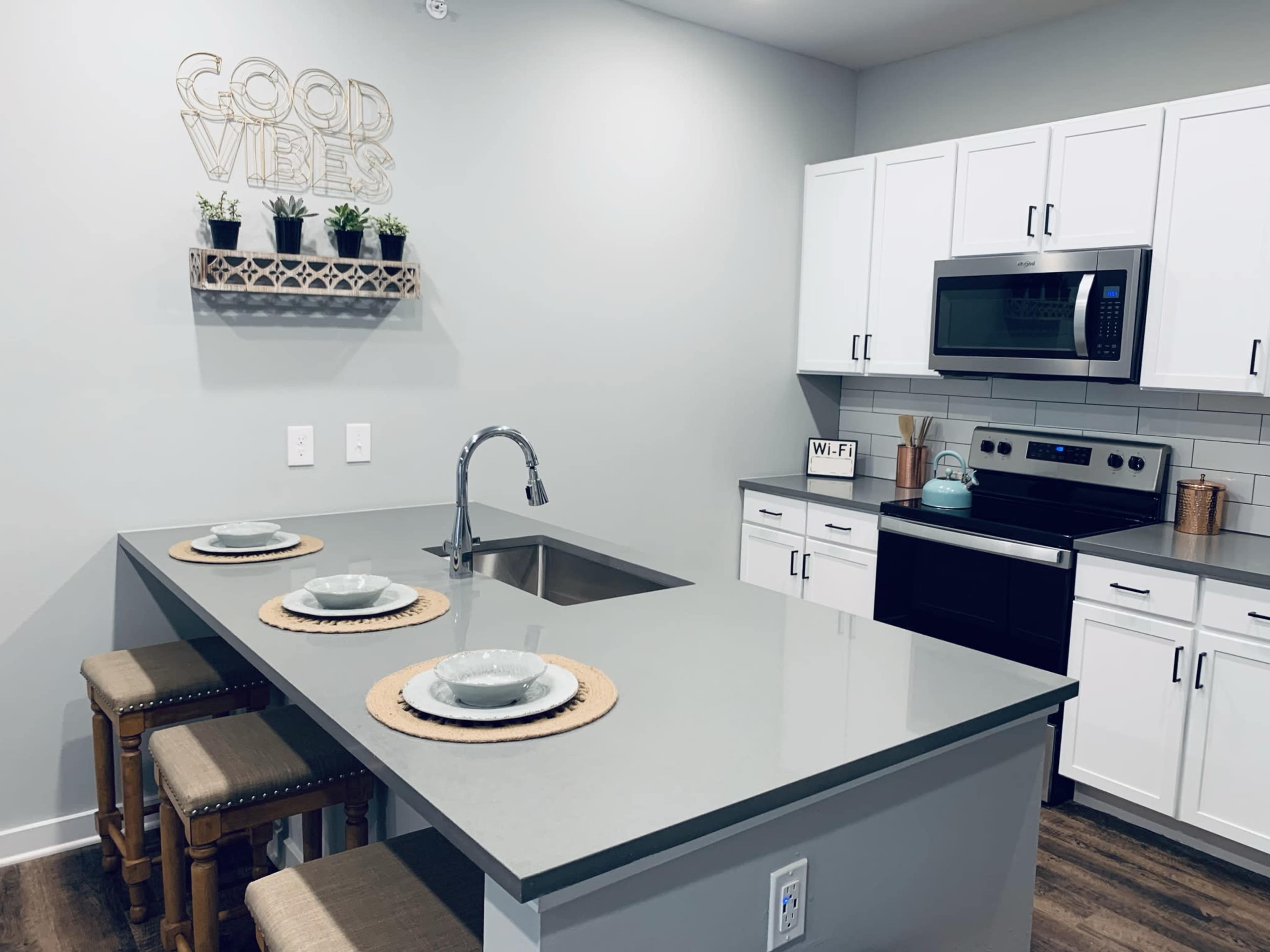 A modern kitchen features a central island with three place settings, stainless steel appliances, and a "Good Vibes" sign on the wall.