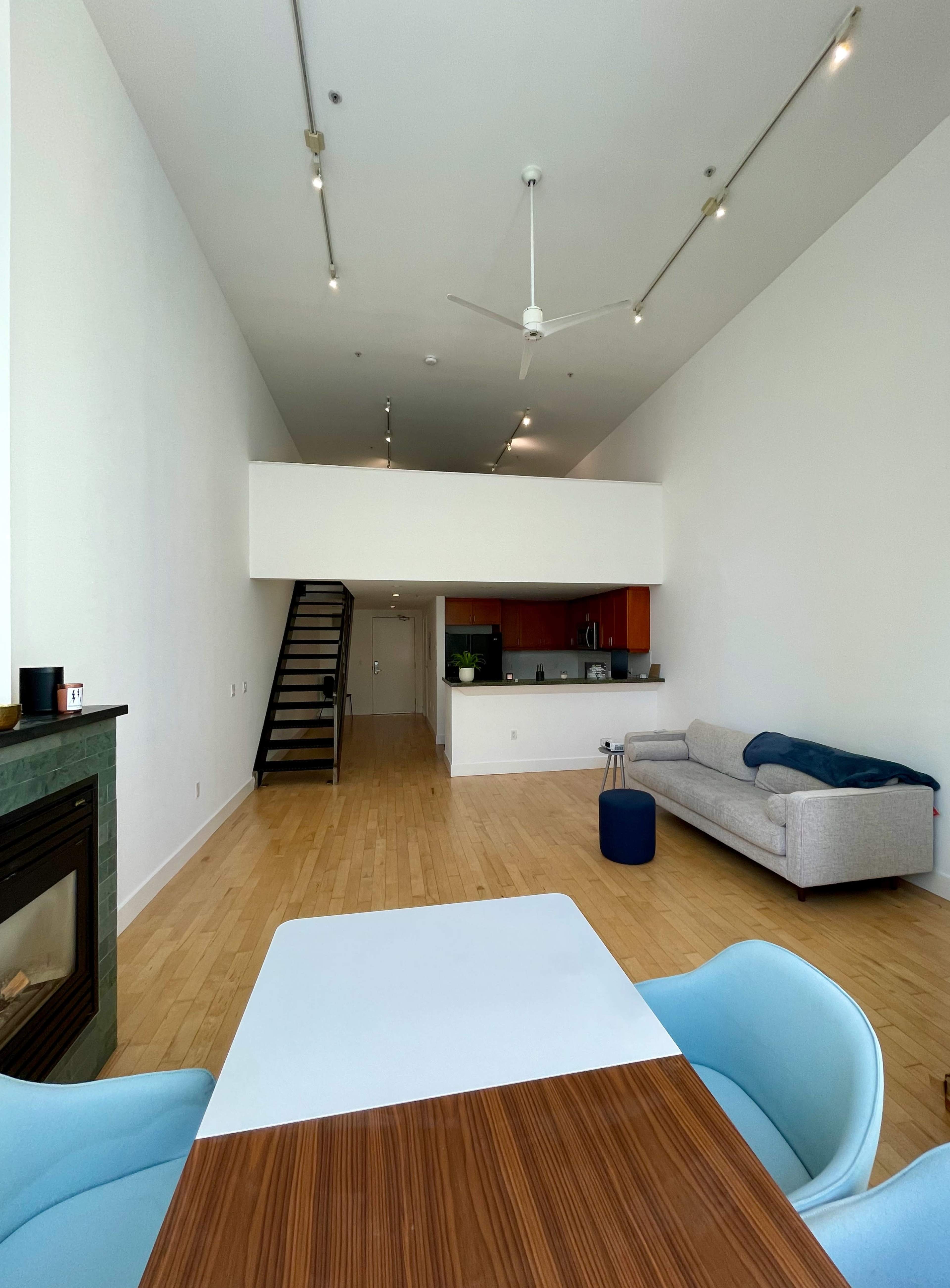 A spacious, modern interior with a dining table in the foreground, a gray sofa along the wall, and a staircase leading to an upper level.