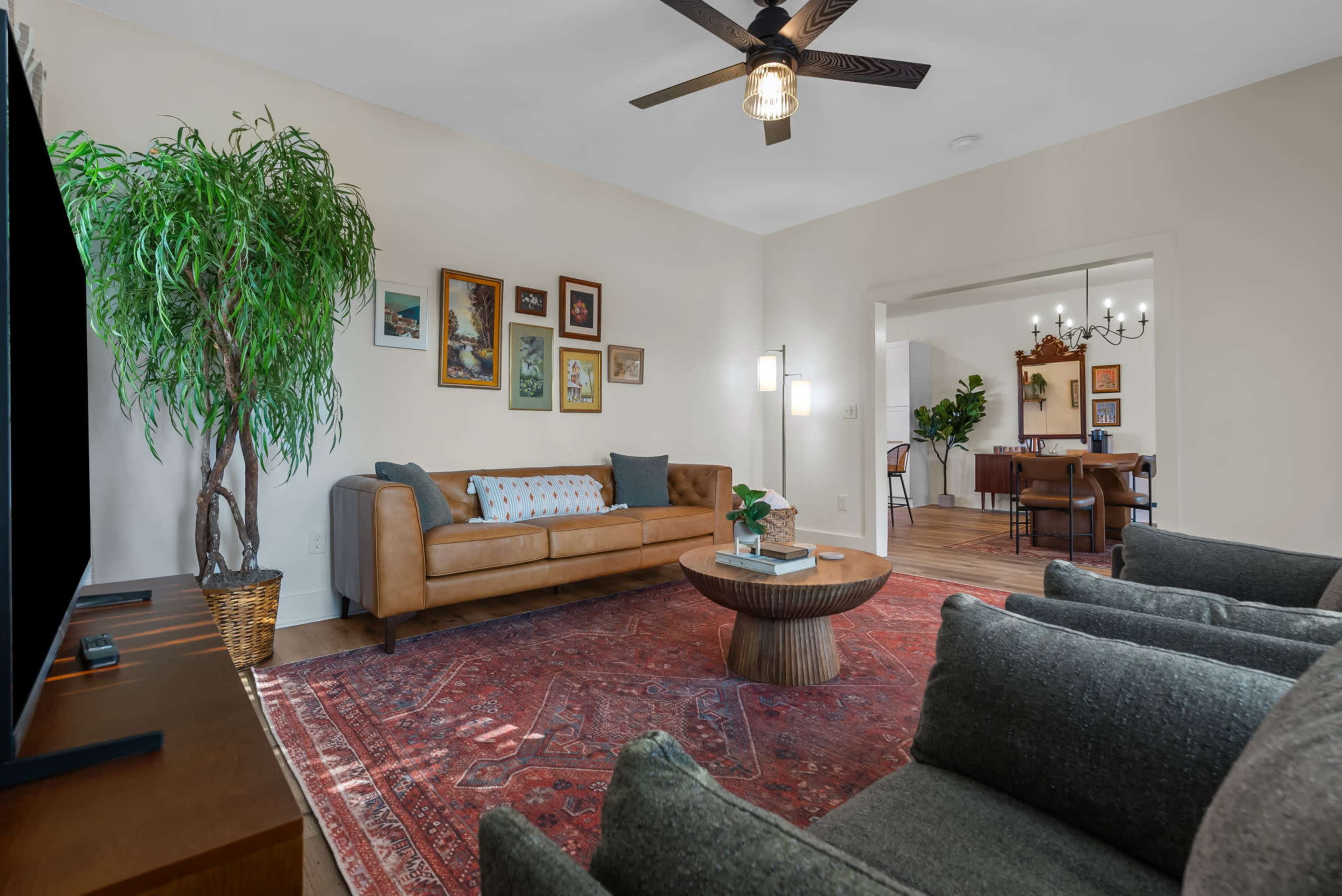 The image shows a living room featuring a brown leather sofa, a central wooden coffee table, green upholstered chairs, a large potted plant, and various framed artworks on the walls.