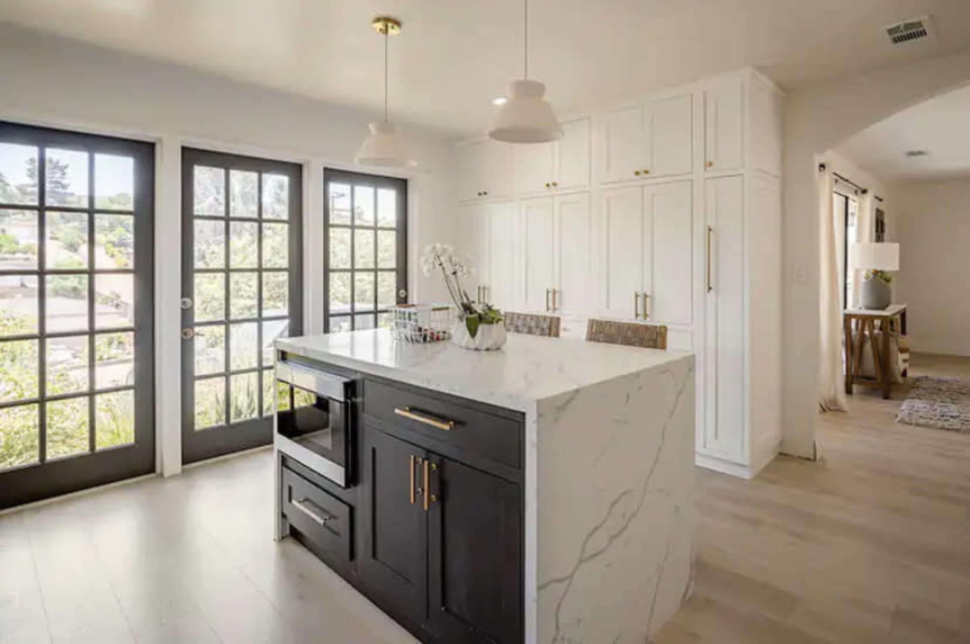 A modern kitchen featuring a central island with a black base and white marble countertop, large windows with black frames, and white cabinetry with gold hardware.