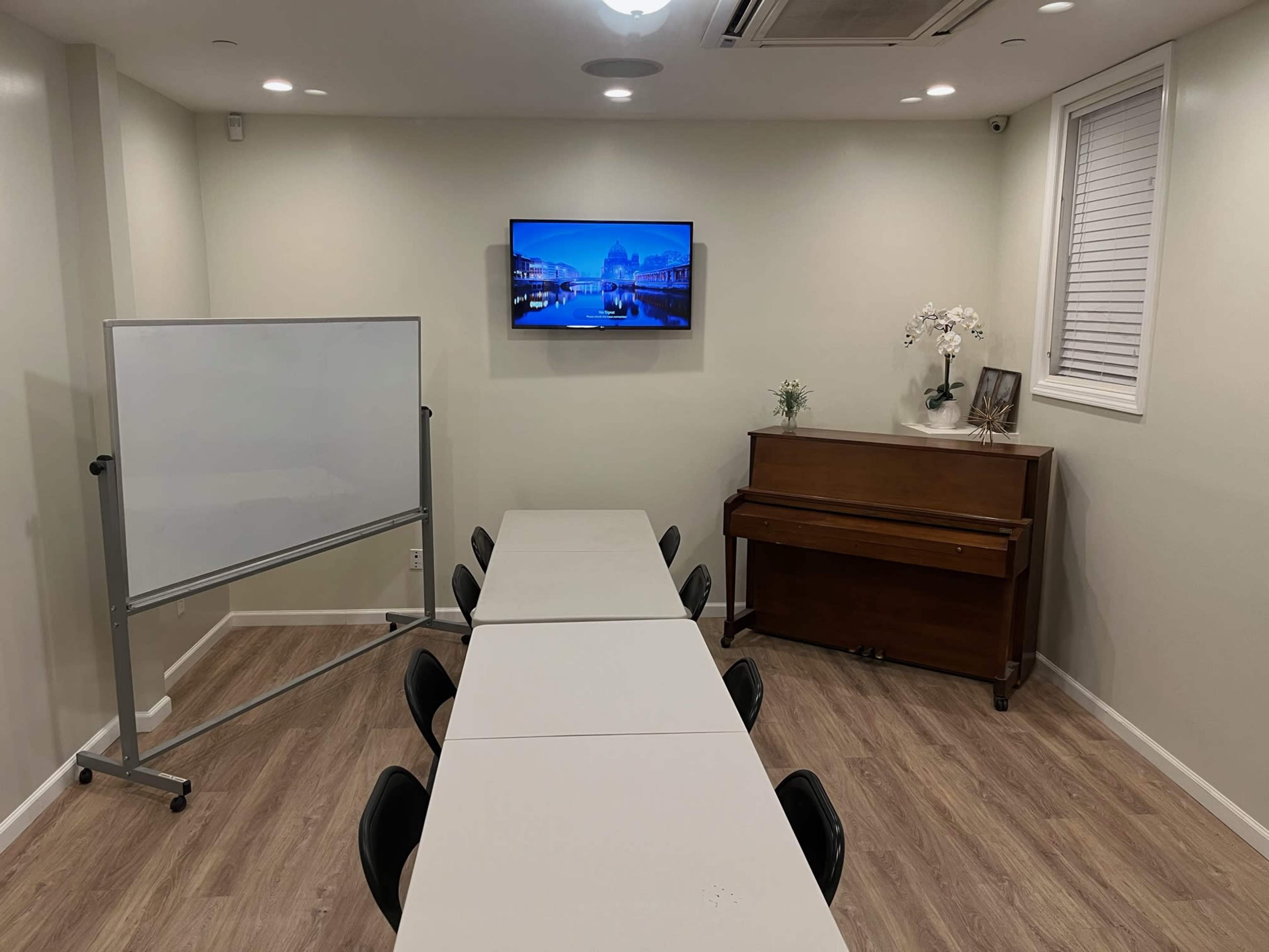 A small meeting room features a rectangular table with black chairs, a whiteboard on one side, and a piano opposite a wall-mounted TV.