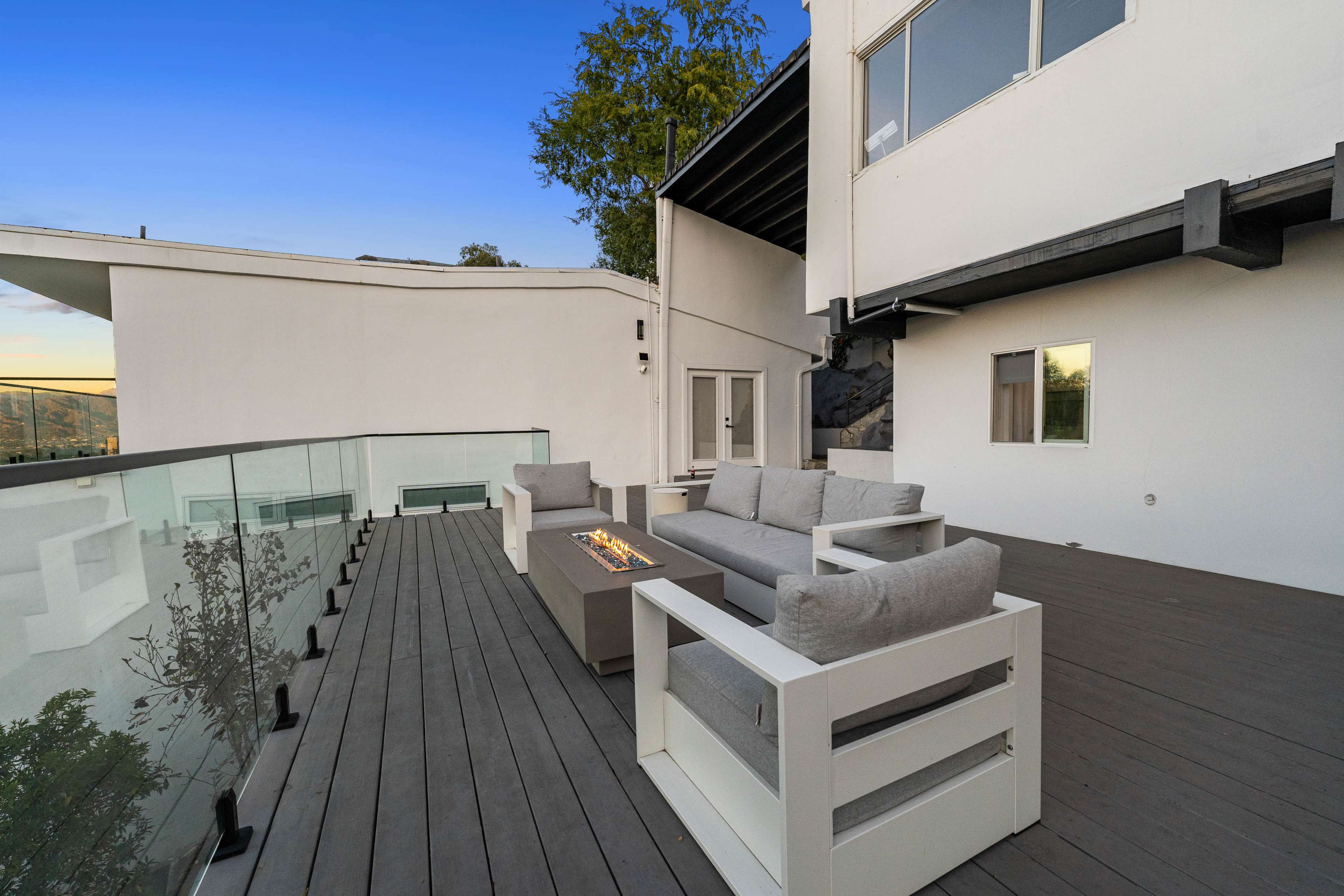 A modern outdoor patio features a seating area with gray cushions, a fire pit, and glass railing overlooking a scenic view.