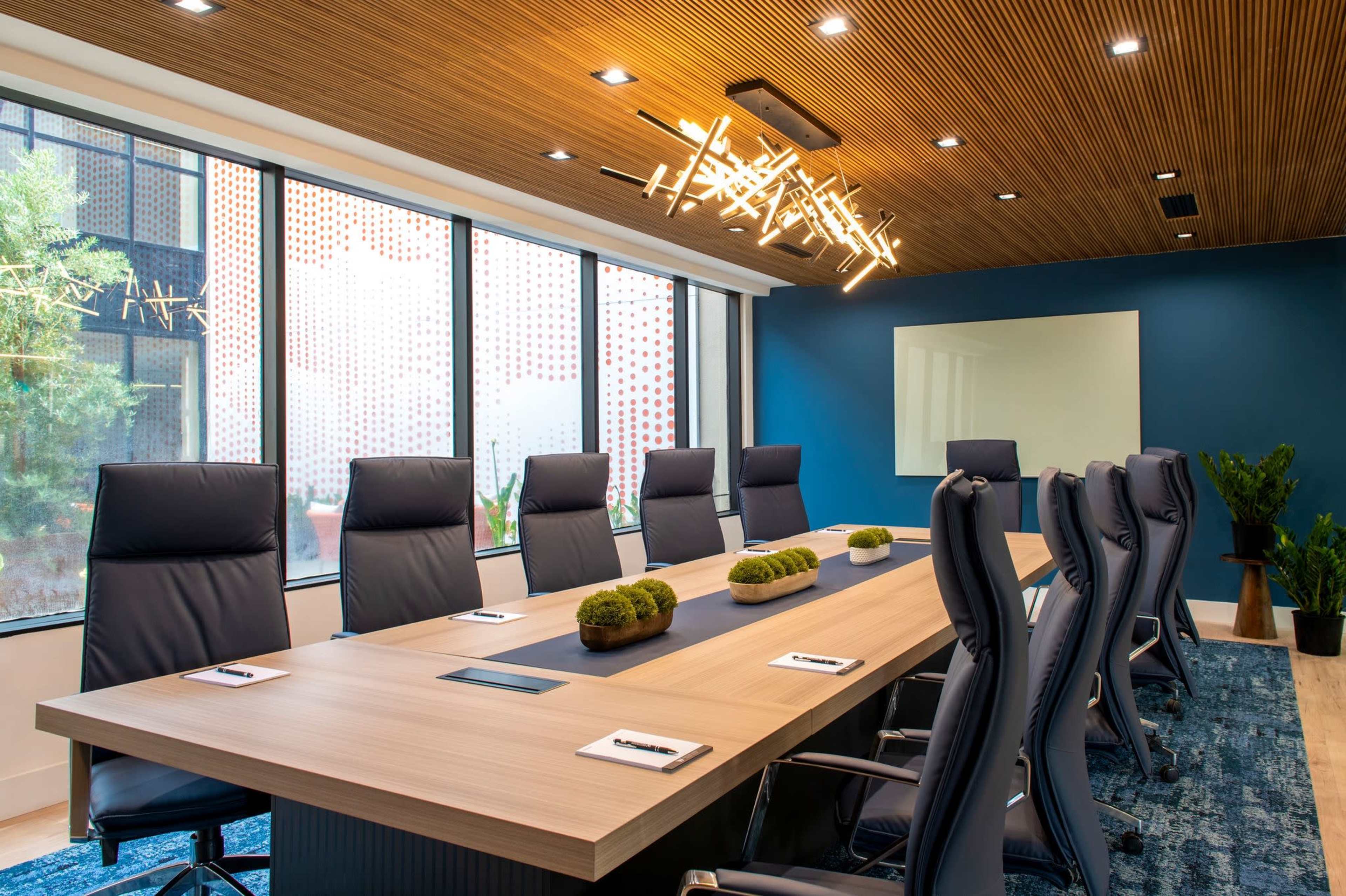 A modern conference room features a large wooden table surrounded by black leather chairs, illuminated by unique overhead lighting and large windows offering a view of greenery outside.