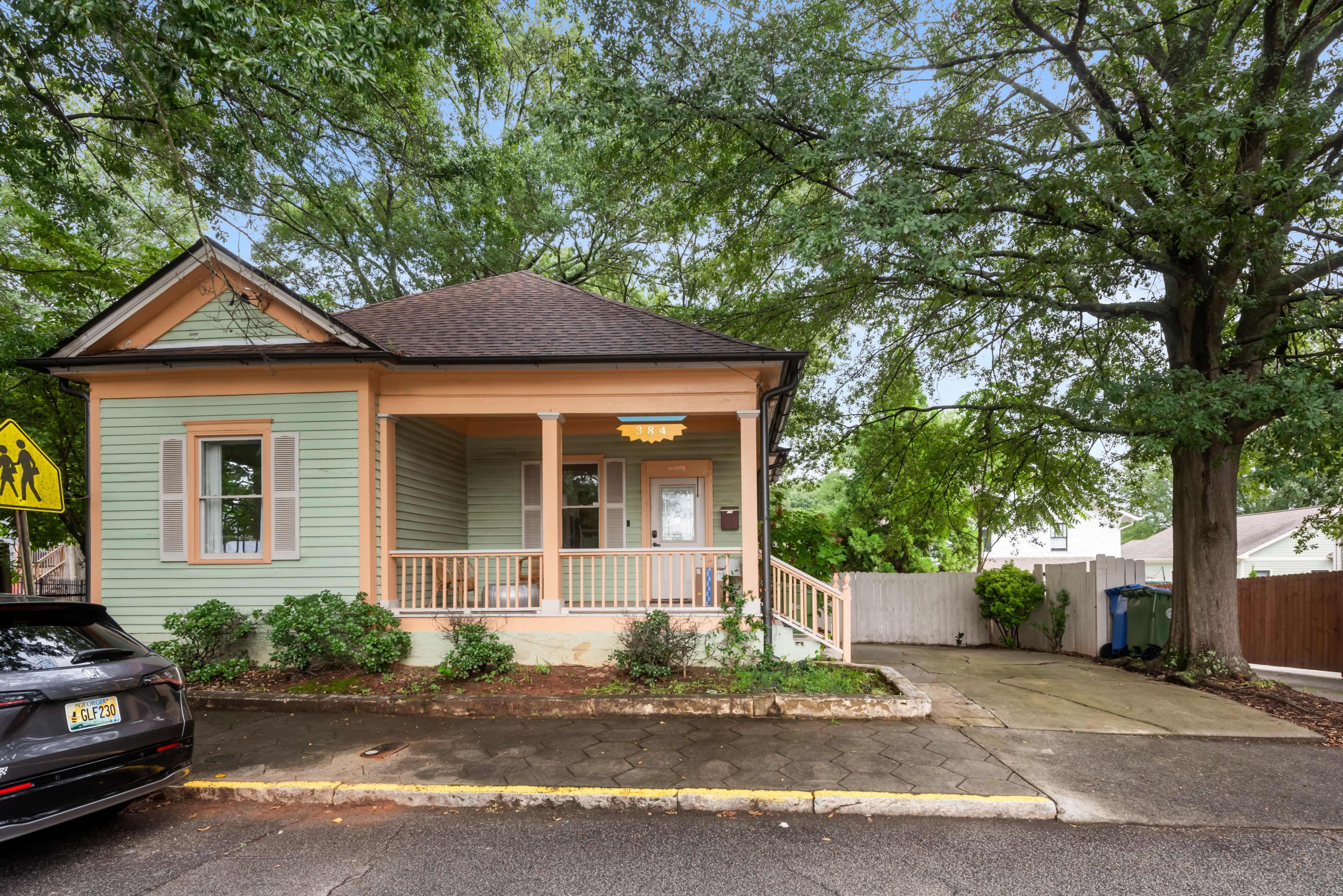 A green wooden house with a porch and a sloped roof sits beside a paved driveway, surrounded by leafy trees.