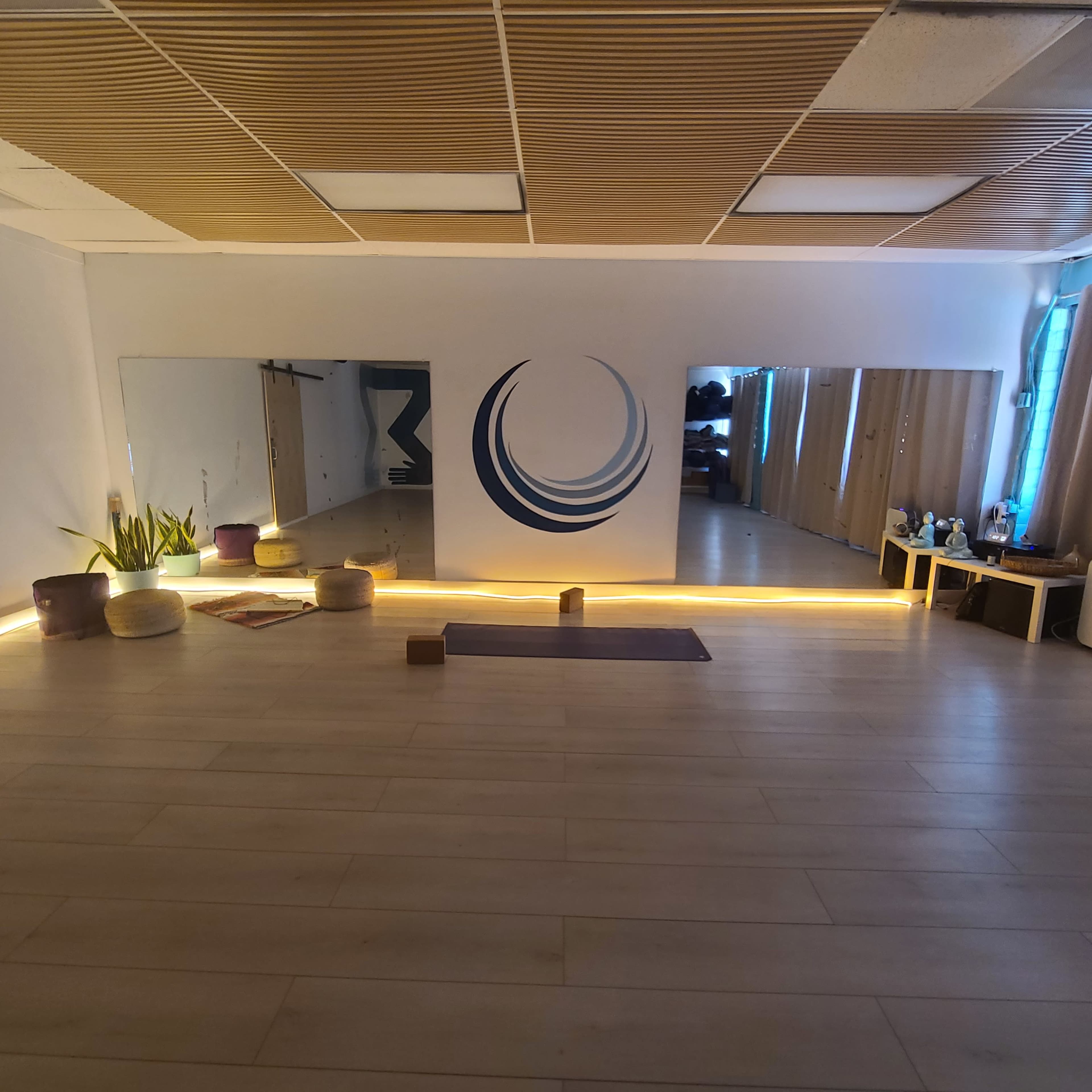 The image shows a yoga studio with wooden flooring, three mirrors on the walls, and a circular design painted on one wall, along with mats and cushions arranged for practice.