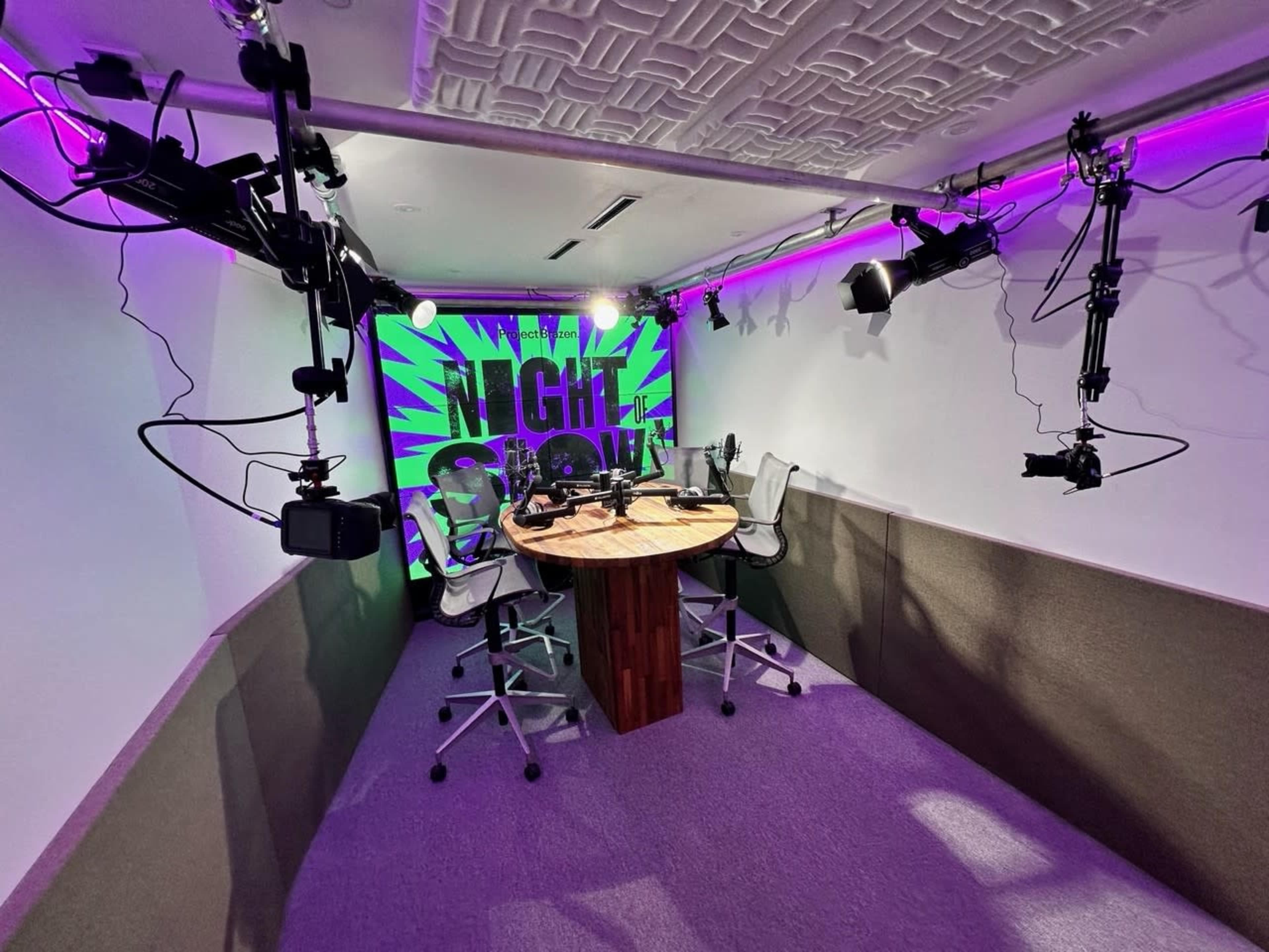 A podcast studio featuring a round table with chairs, surrounded by cameras and vibrant purple lighting on the walls.