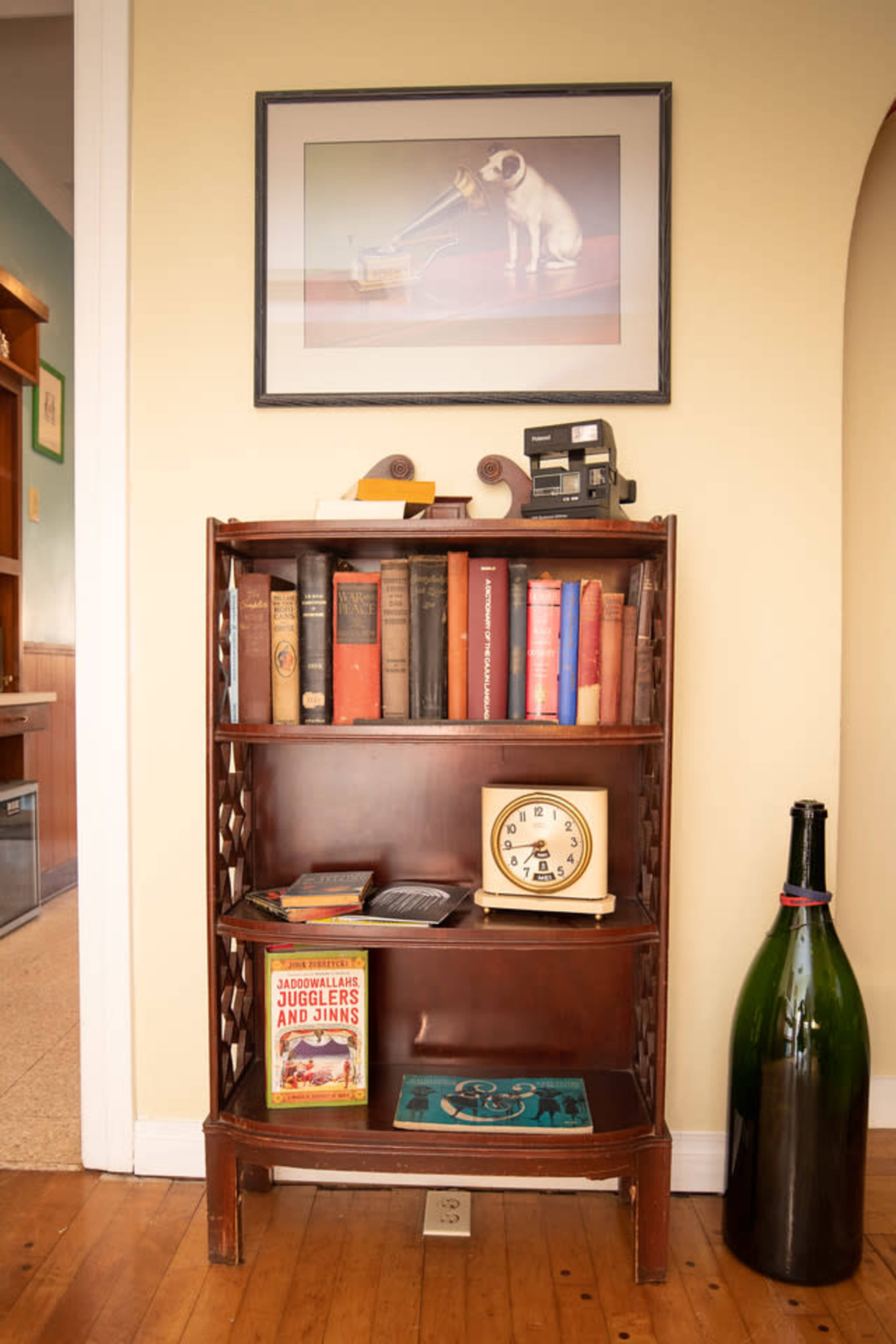 A wooden bookshelf filled with books, a clock, and a large wine bottle stands against a wall featuring a framed artwork of a dog.