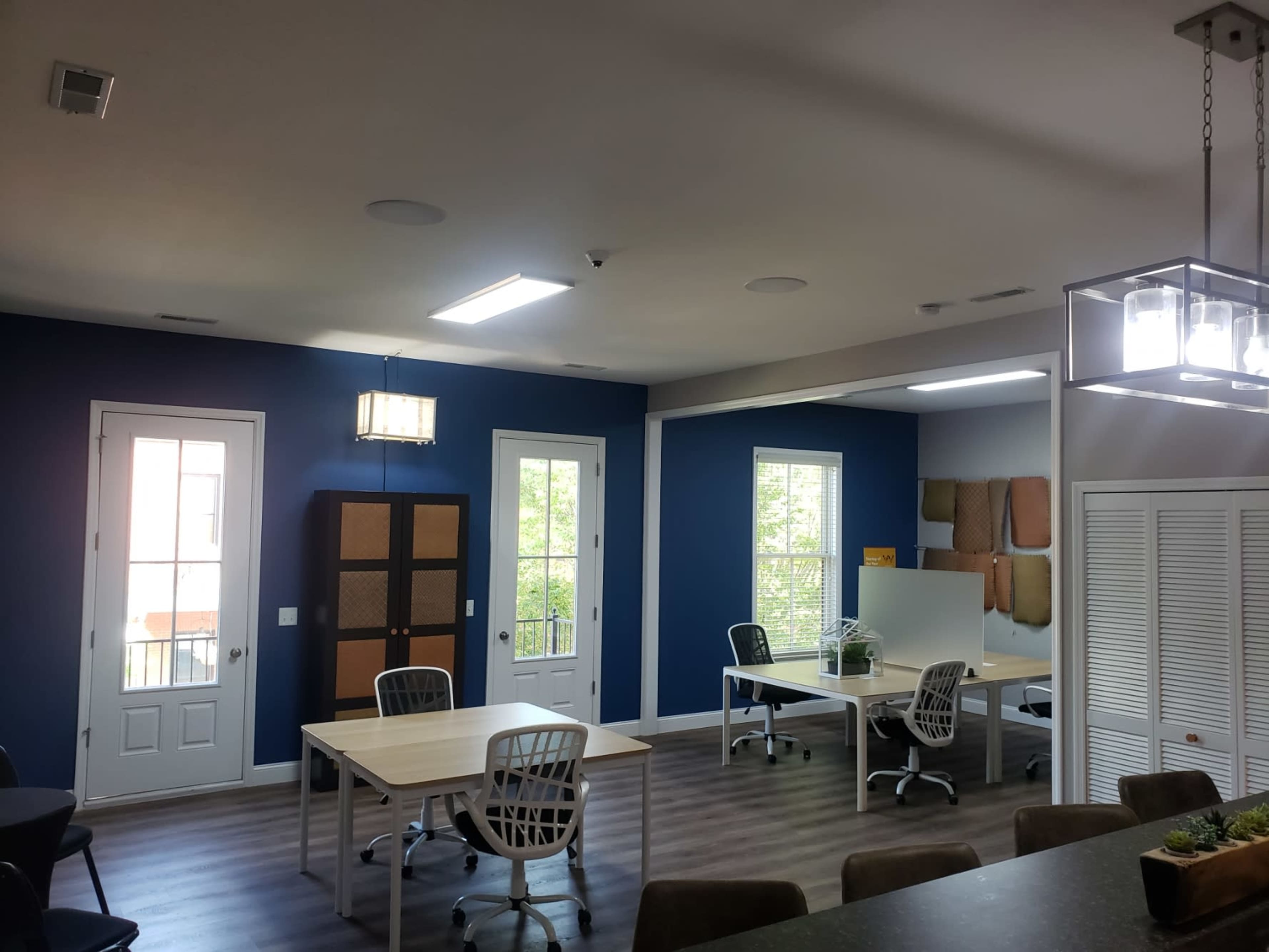 A modern office space with several wooden desks and chairs, blue walls, and large windows allowing natural light to enter.