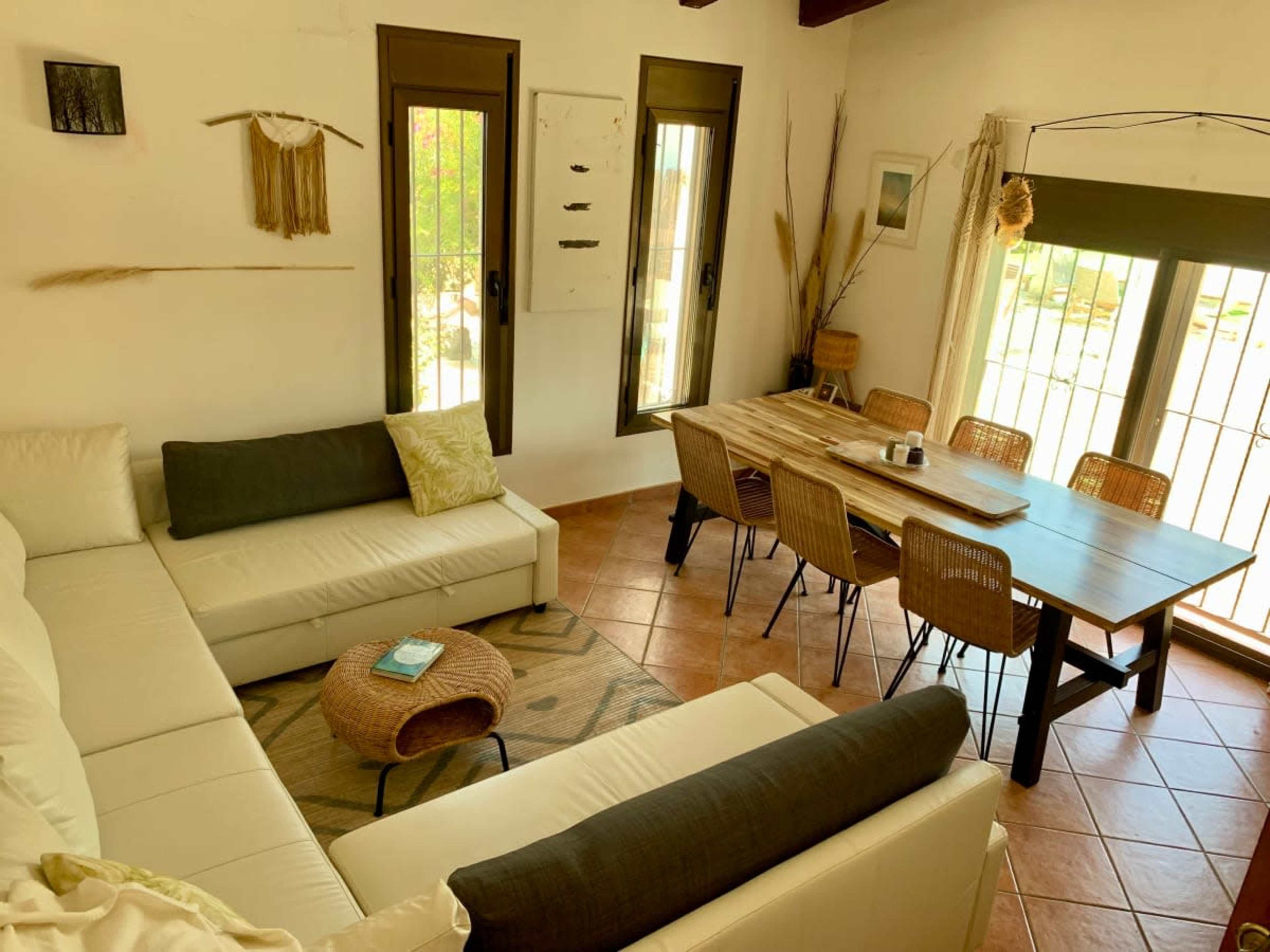 The image shows a bright living space featuring a white sectional sofa, a wooden dining table with chairs, and large windows allowing natural light to fill the room.