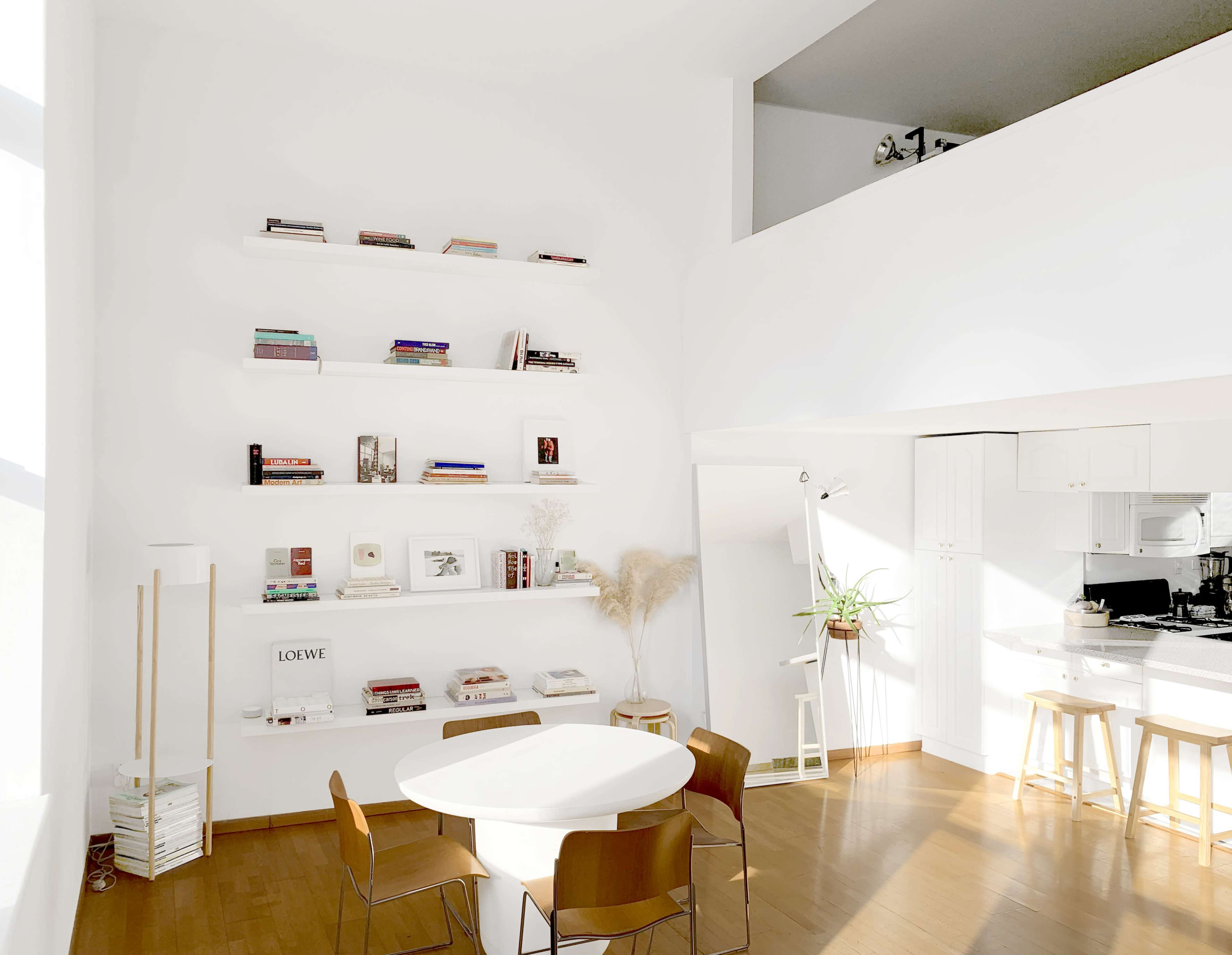 The image shows a bright, minimalistic living space with a circular table surrounded by wooden chairs, a tall shelf displaying books, and a kitchenette in the background.