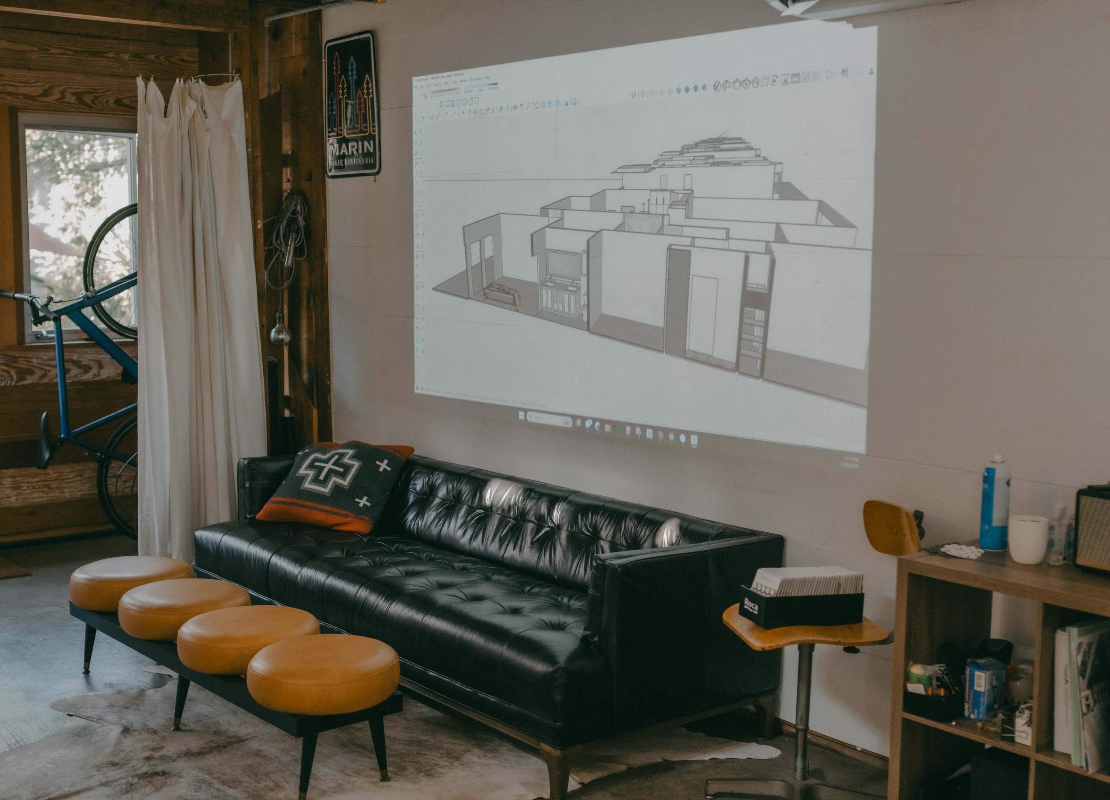 A black couch with orange cushions is positioned in front of a wall where a 3D architectural design is projected.