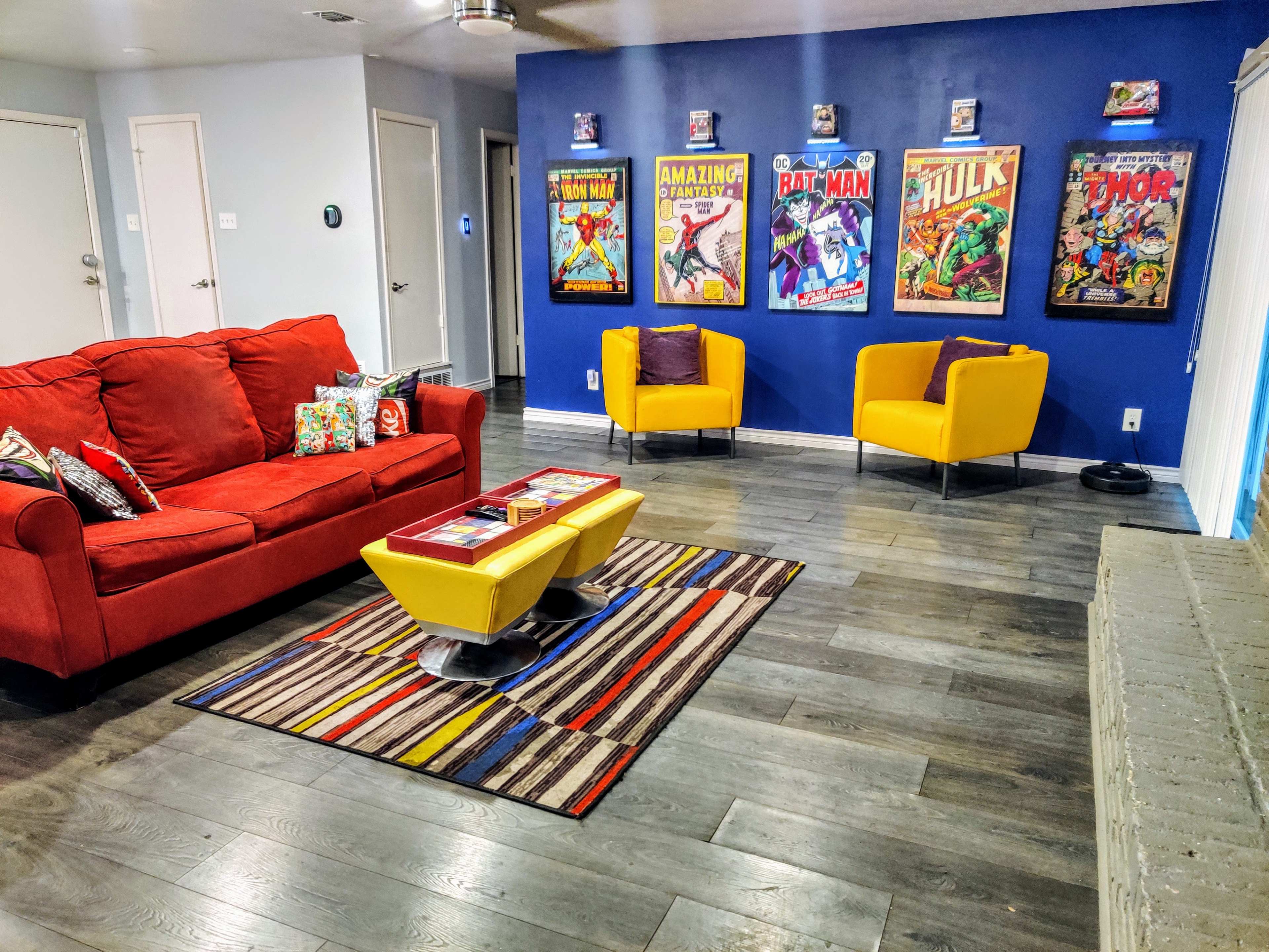 A brightly decorated living room features a red couch, yellow chairs, a striped rug, and framed comic book posters on the blue walls.