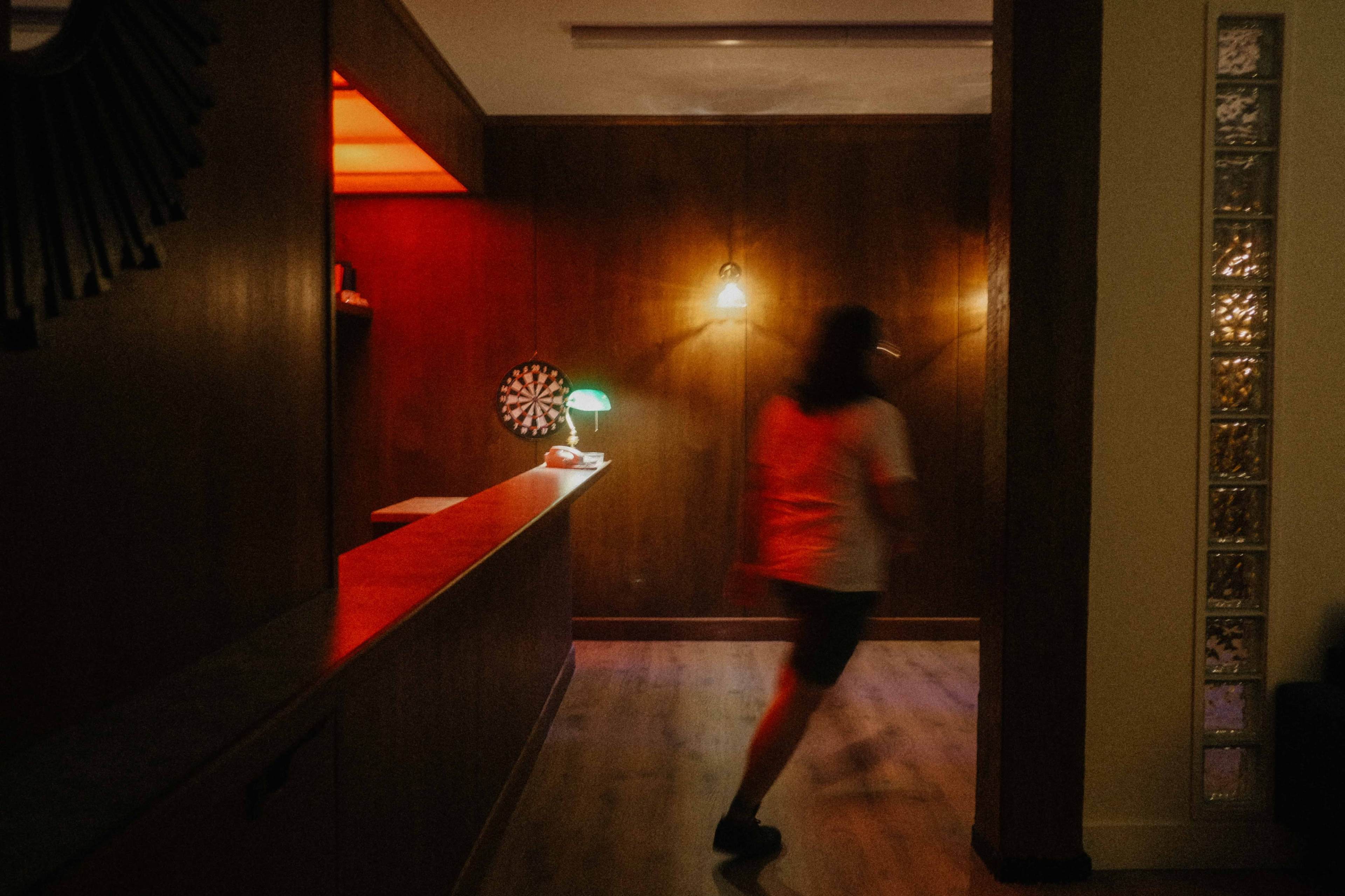 A person walks through a dimly lit room with wooden walls and a dartboard on the side.